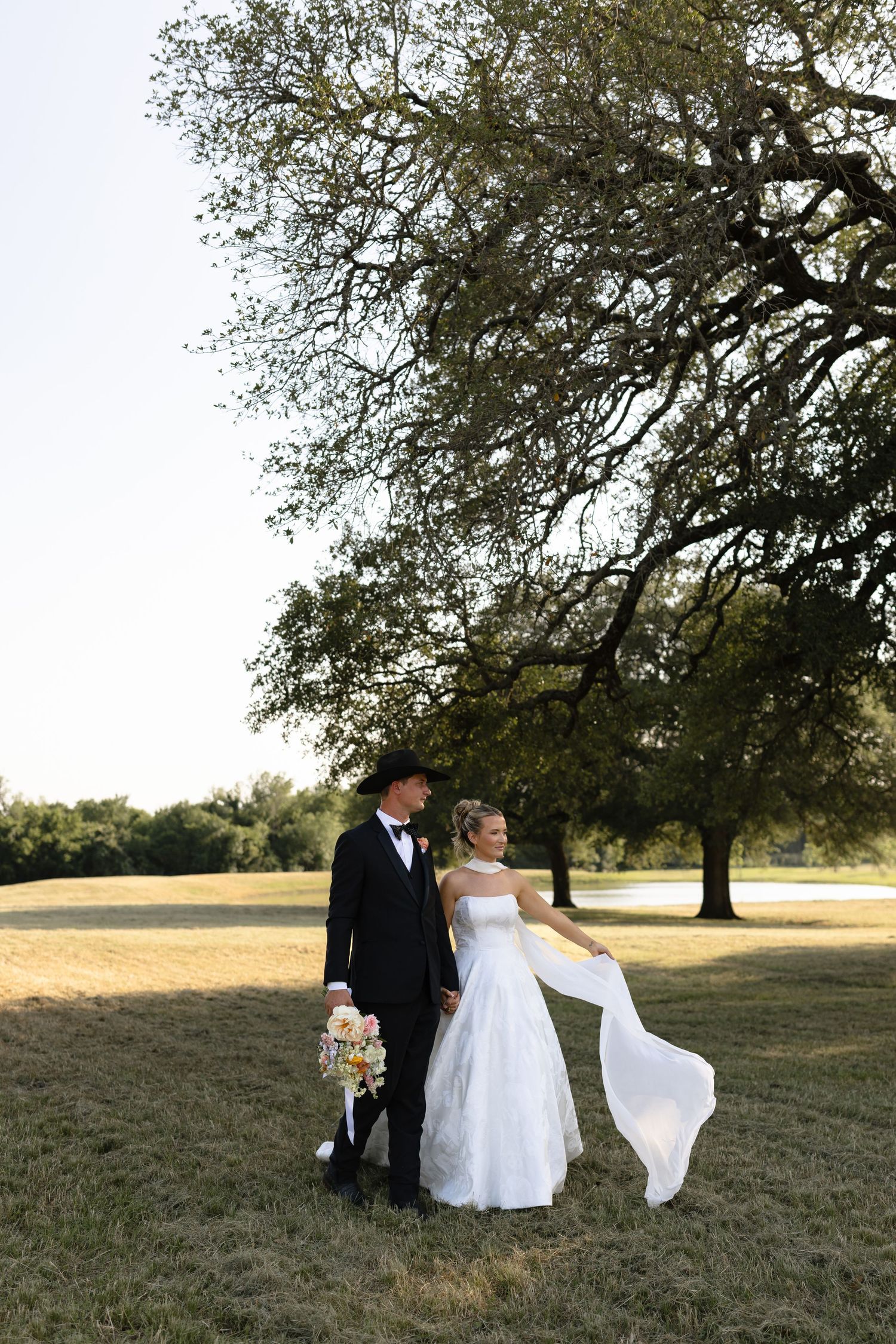 A bride in a white flowing dress and groom in a black tuxedo stand beneath a large tree in a grassy field.