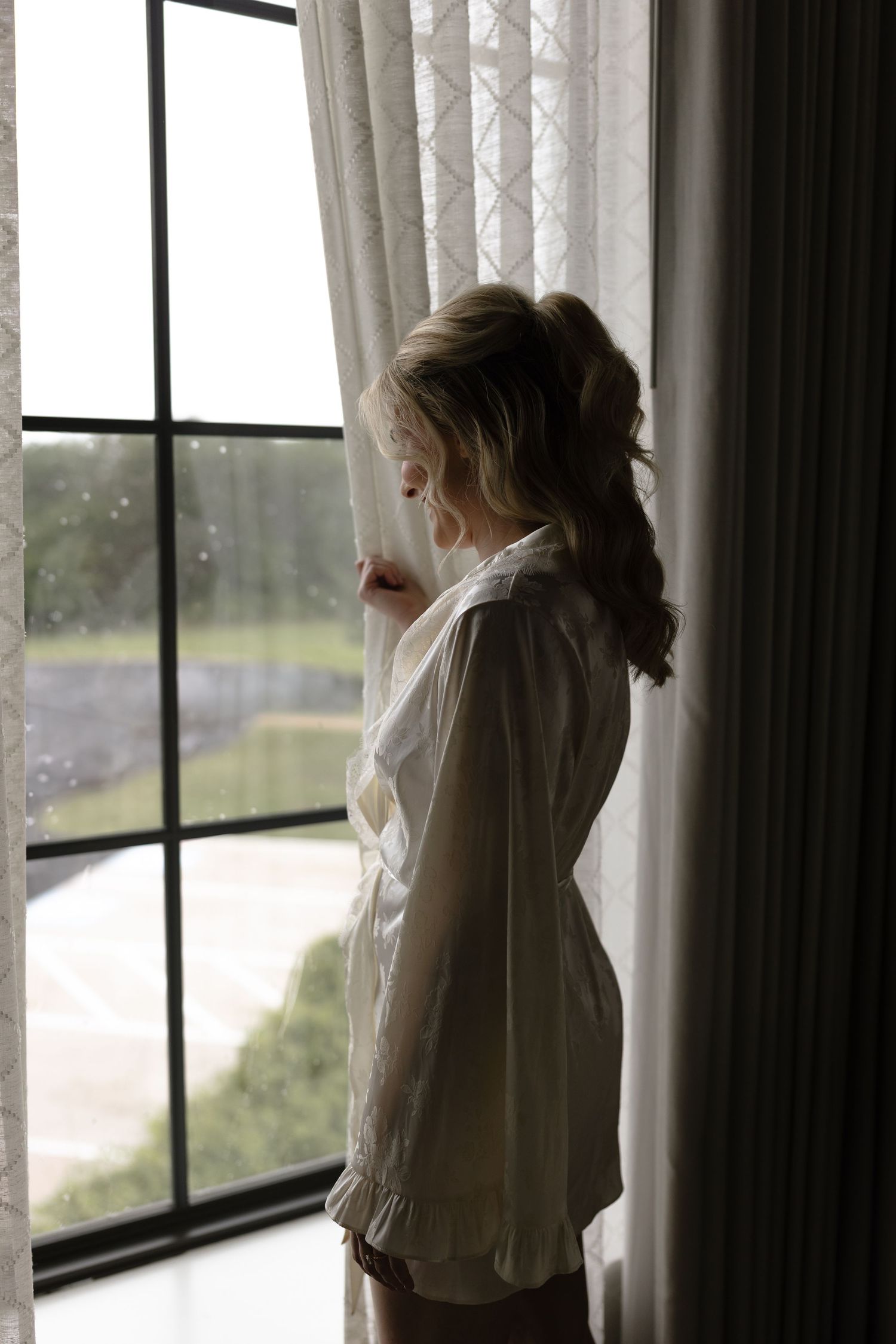 Figure in white garment gazes through window with sheer curtains in moody natural lighting.