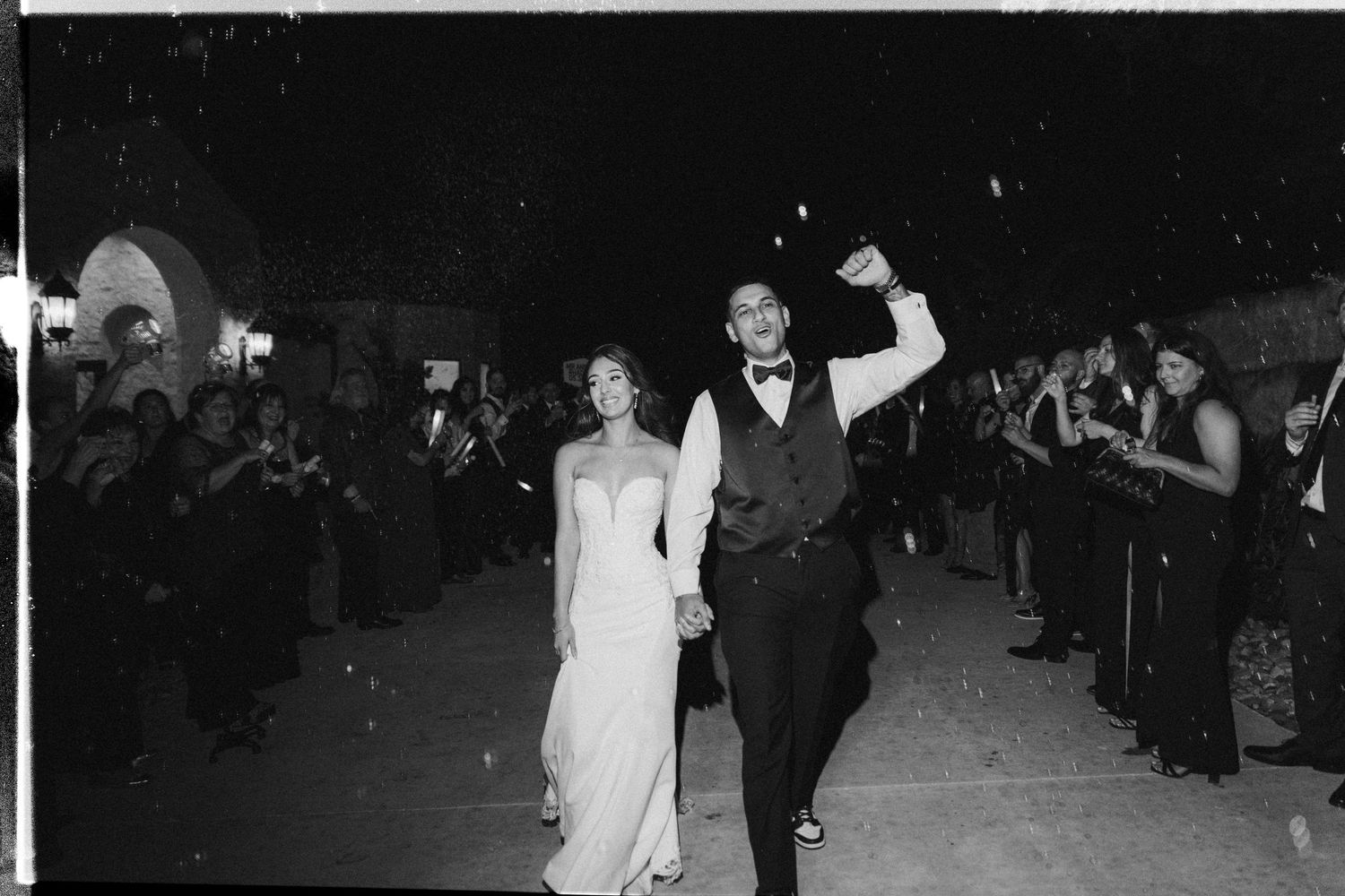 Black and white photo of a bride and groom making their wedding exit while guests line the walkway at night.