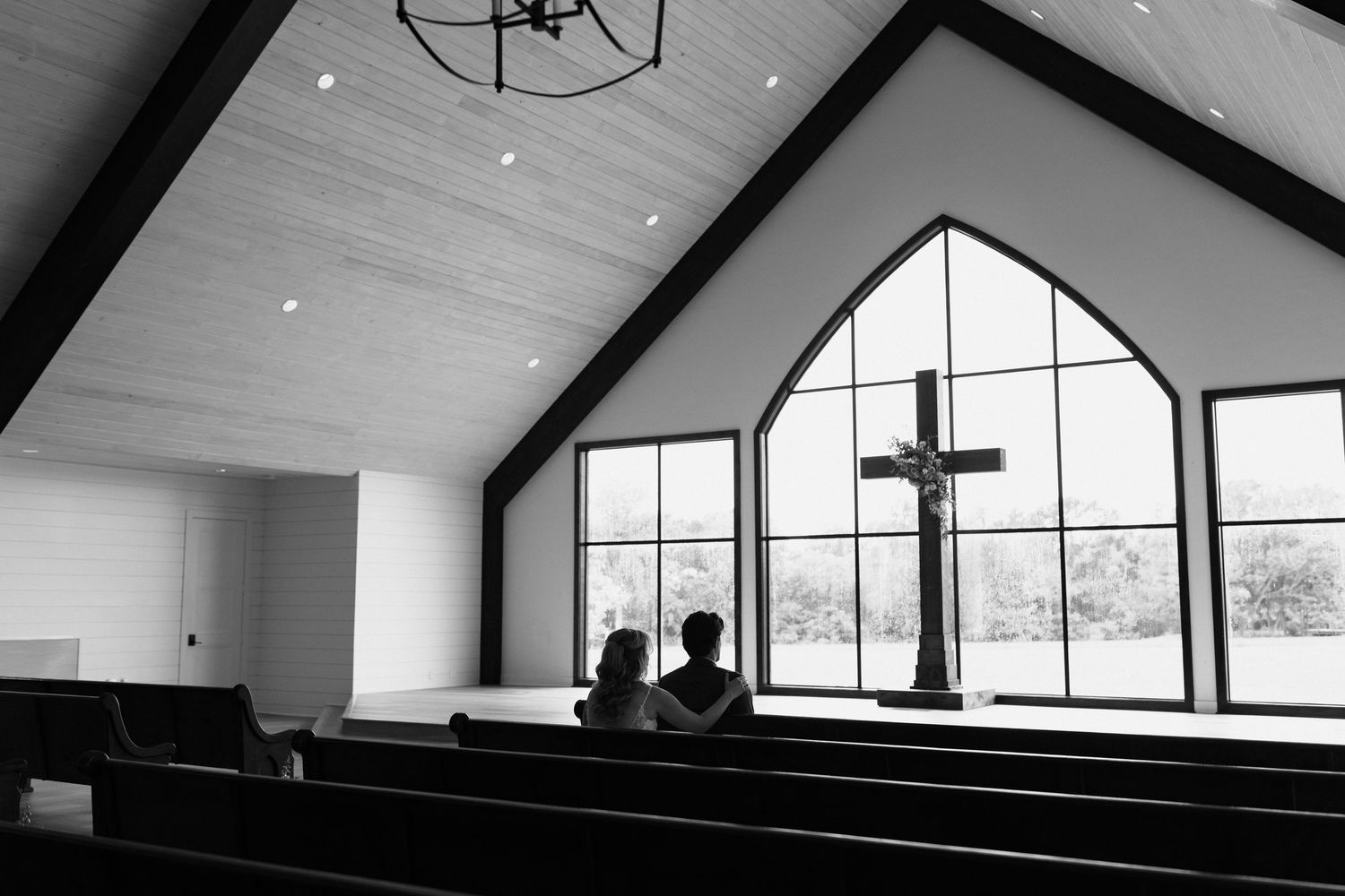 Two silhouetted figures sit in wooden pews inside a modern church with large arched windows and a cross at the altar.