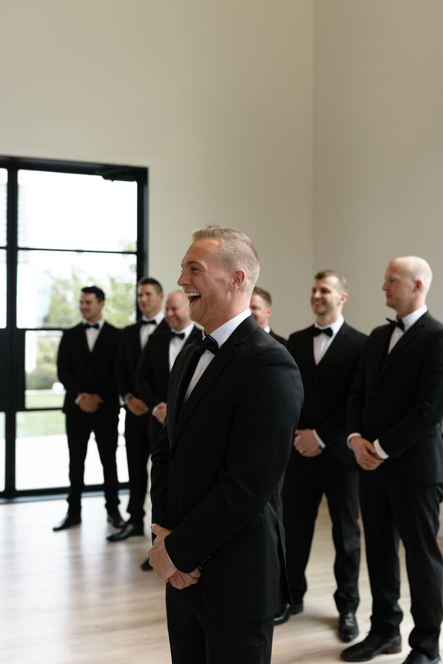 Groomsmen dressed in formal black tuxedos stand in line during wedding preparation in a bright modern venue.