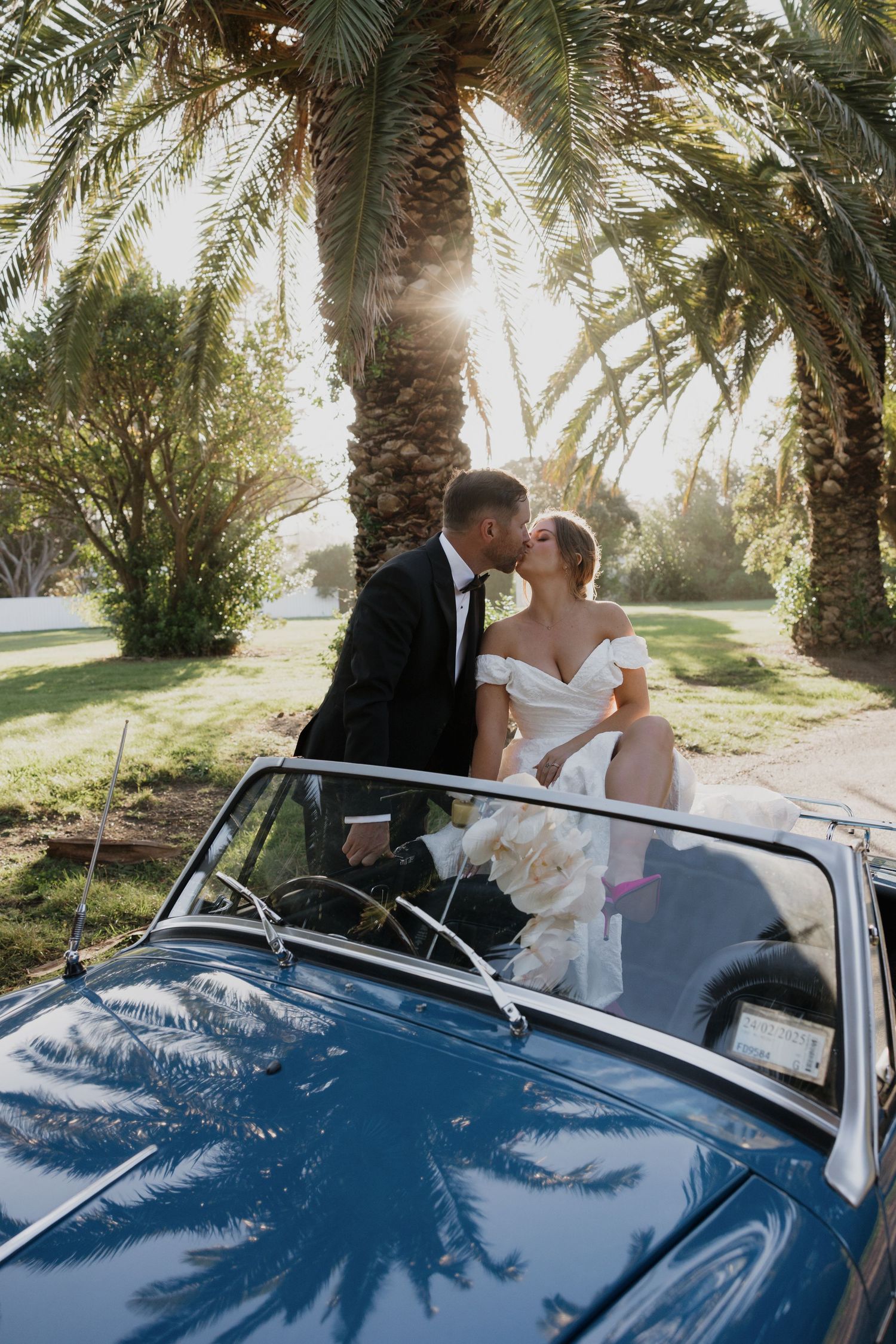Newlyweds share a romantic moment in a vintage convertible car under palm trees.