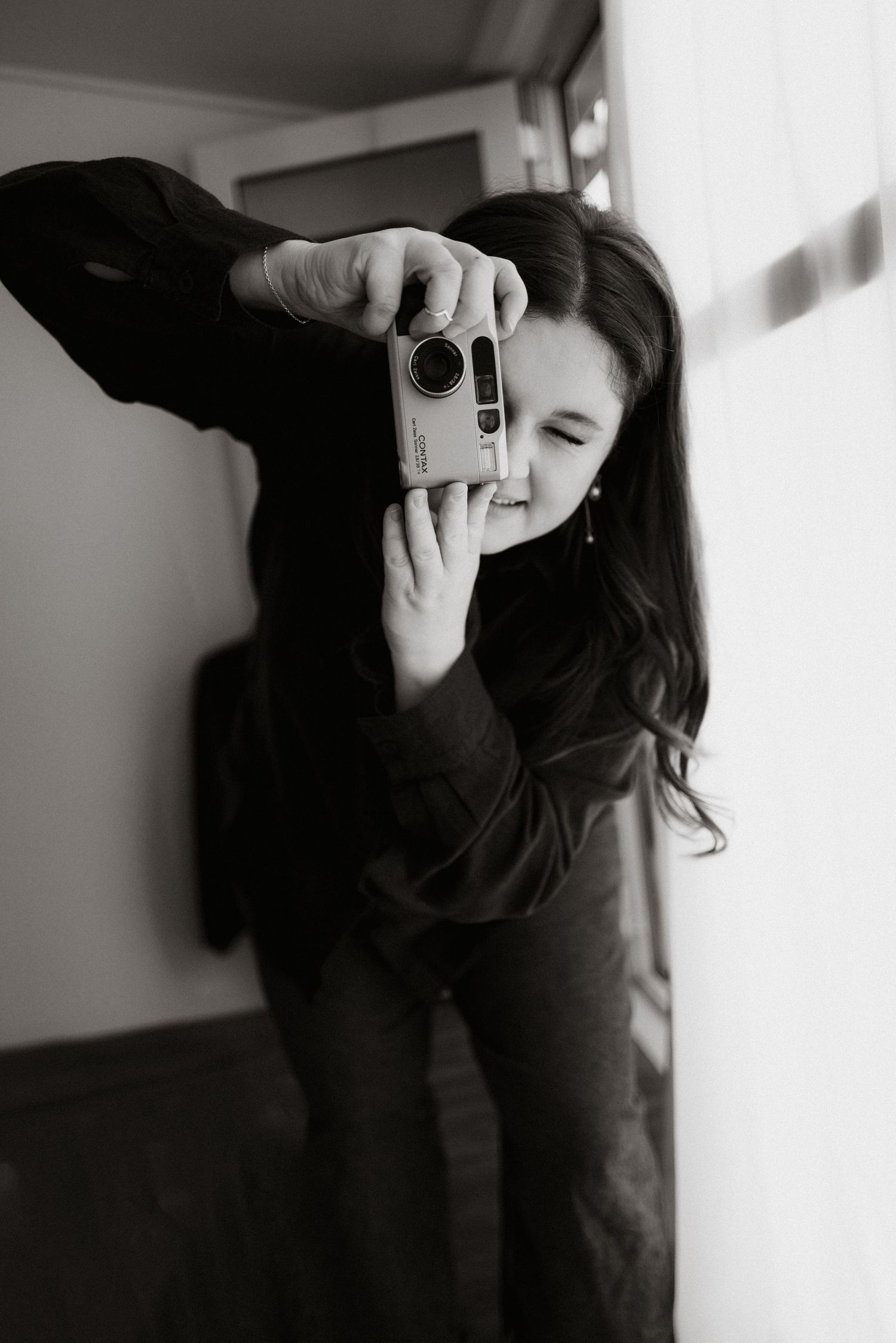 Black and white photo of someone taking a picture with a compact camera in an indoor setting.