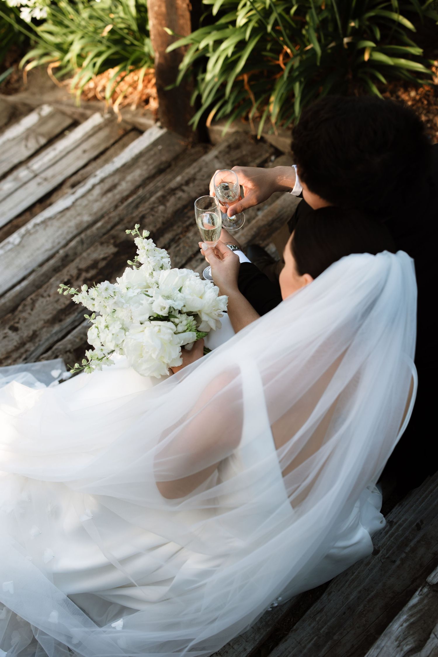A bride in flowing white dress holds a drink next to white floral arrangements.