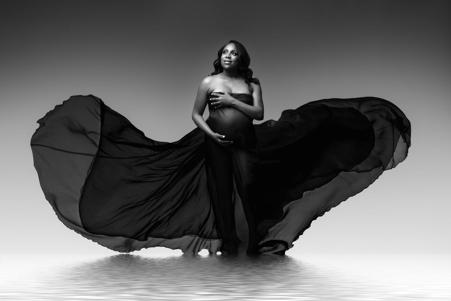 Artistic black and white maternity photo with billowing fabric creating dramatic movement.