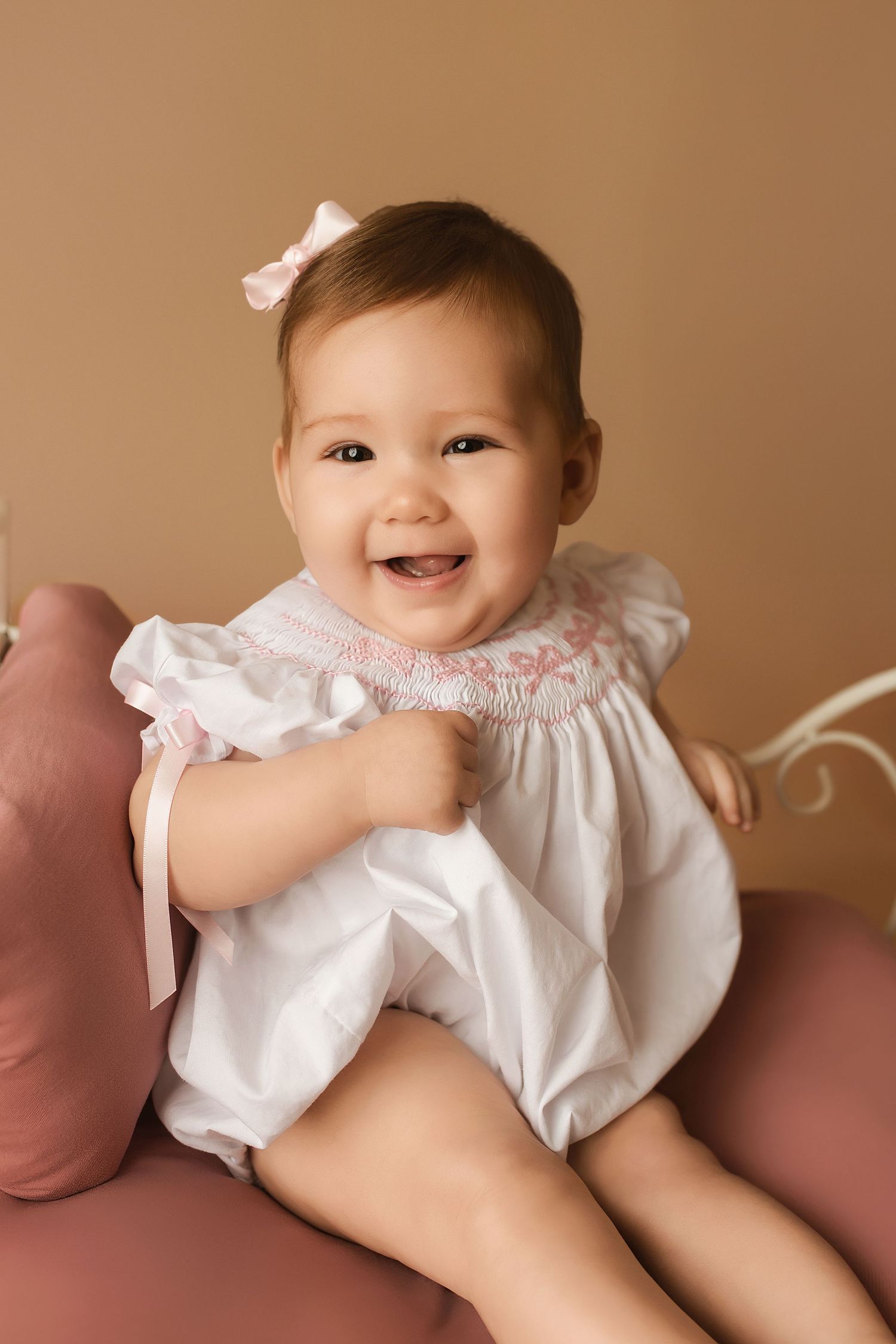 Baby wearing white dress sitting on pink cushions during professional photo session.
