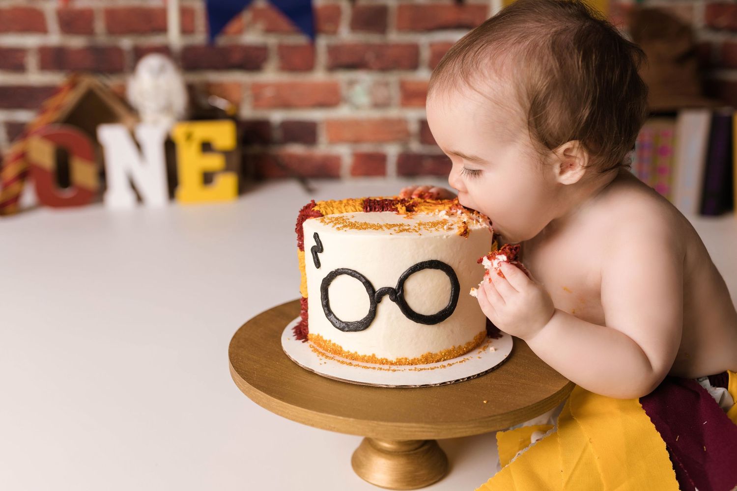 Baby near birthday cake with glasses decoration on brick wall background.