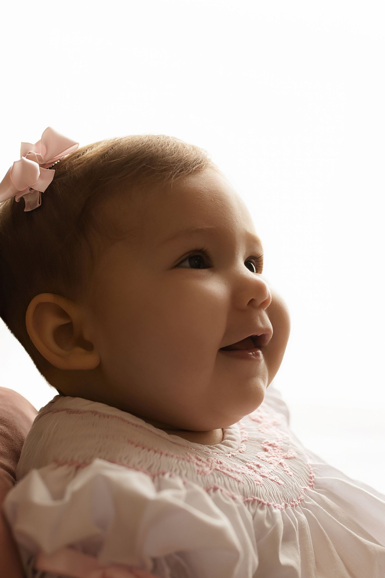 Baby in delicate pink smocked dress gazes upward against white background.