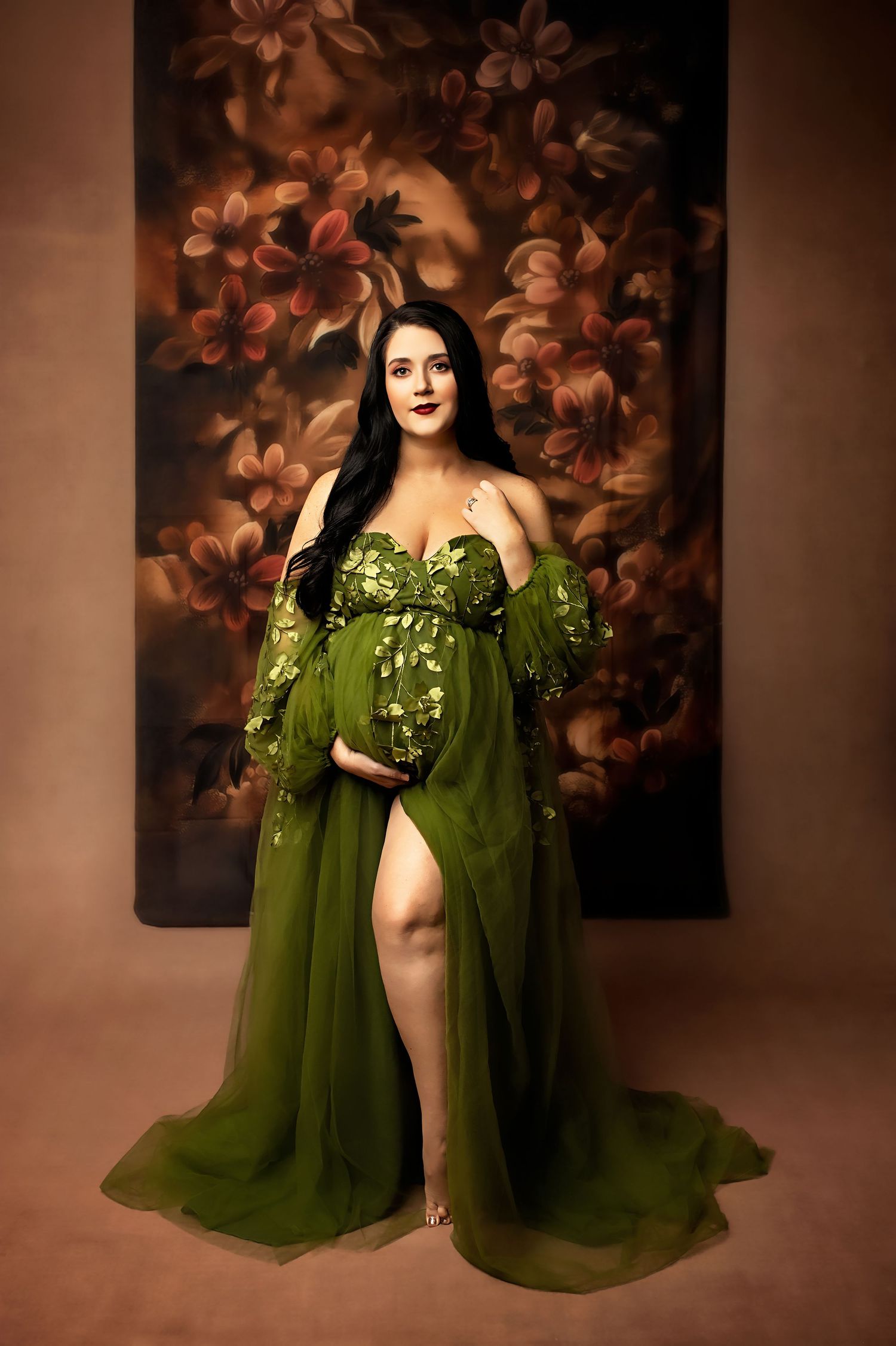 Dramatic maternity portrait in flowing olive green gown against warm floral backdrop.