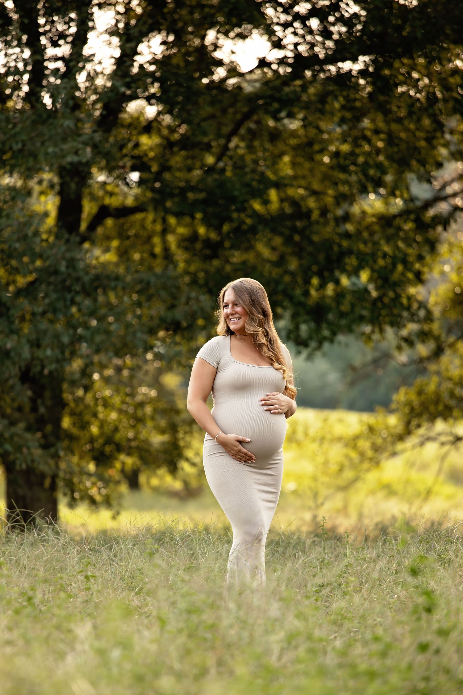 Expectant mother poses in a field at golden hour wearing a fitted white dress.