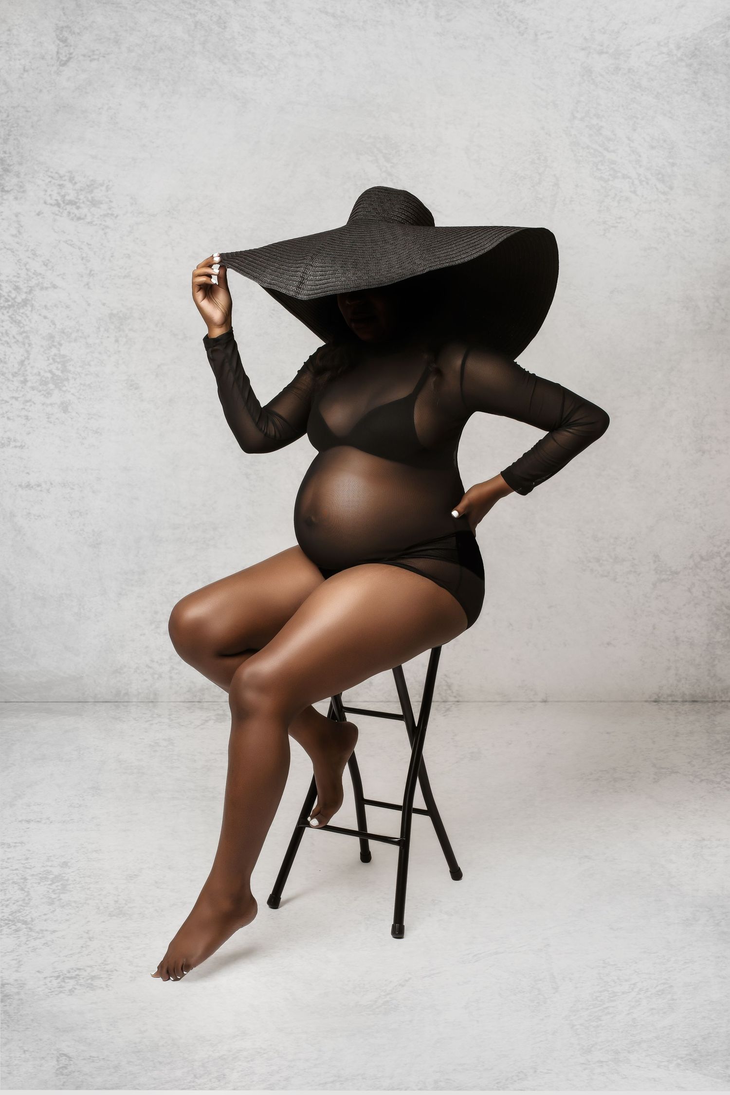 Stylish maternity portrait with wide-brimmed hat and sheer black dress on stool.