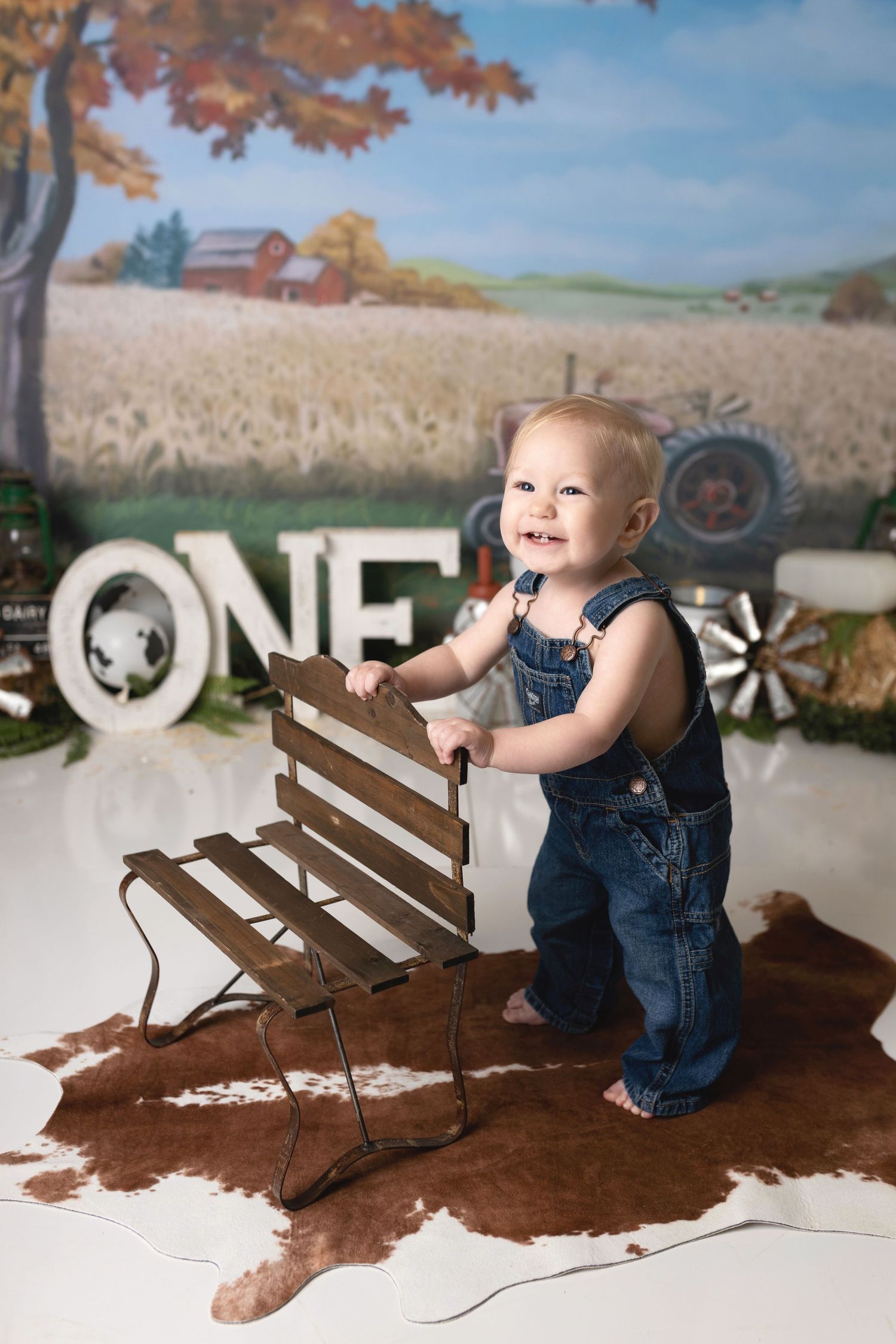 Baby in overalls playing on rustic farm set with autumn backdrop.