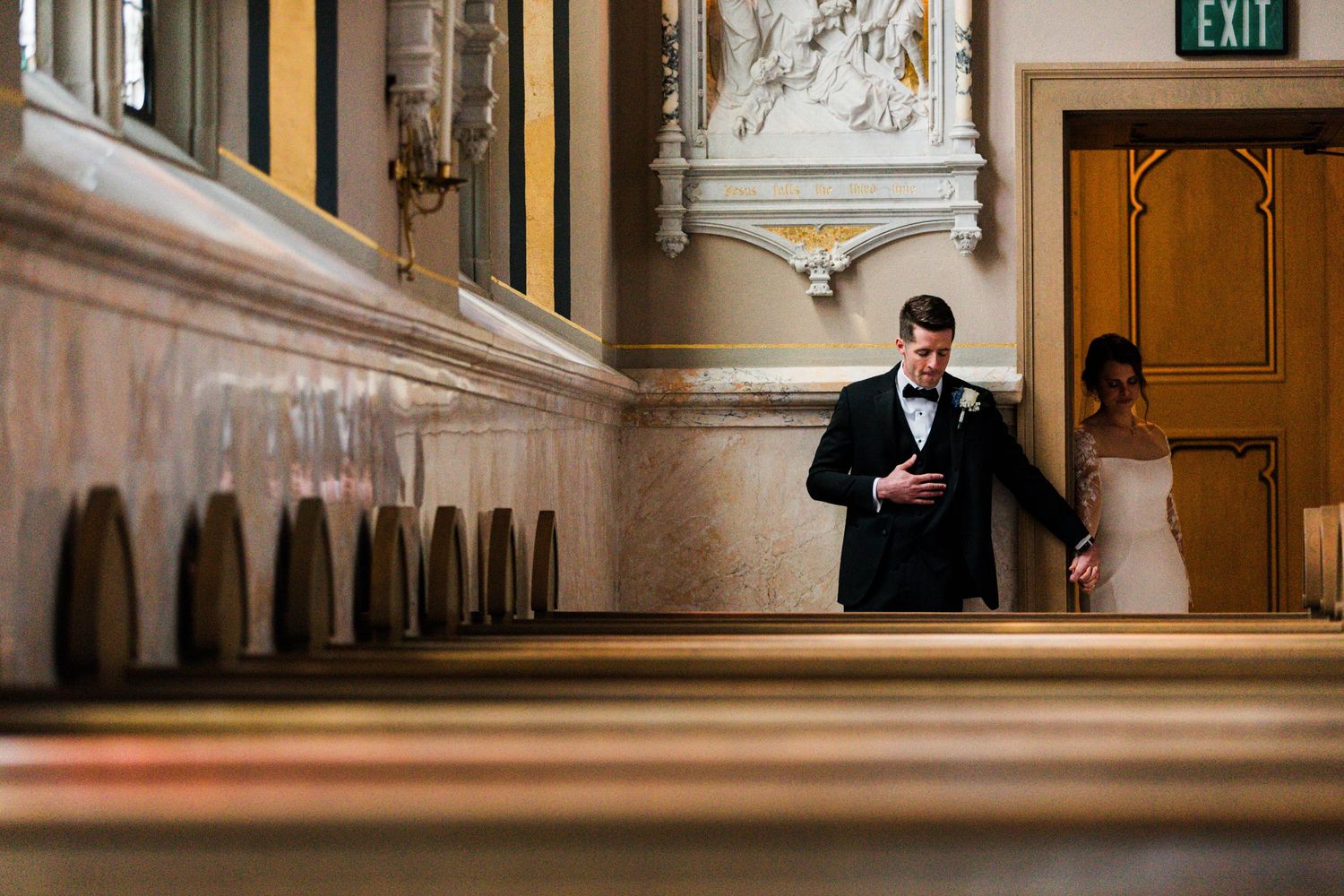 Wedding couple walking through ornate church hallway with decorative architecture and exit sign.
