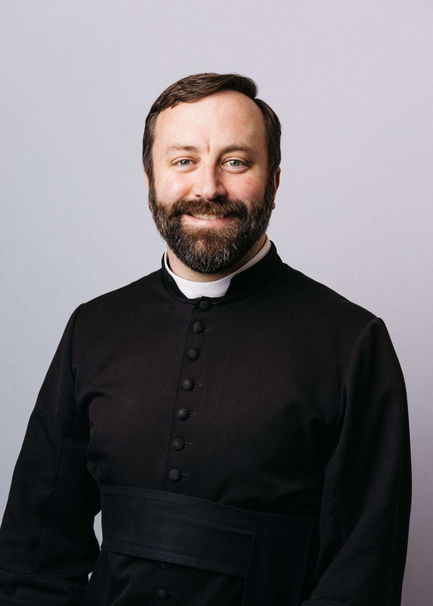 Person wearing traditional black clerical attire with collar against gray background.