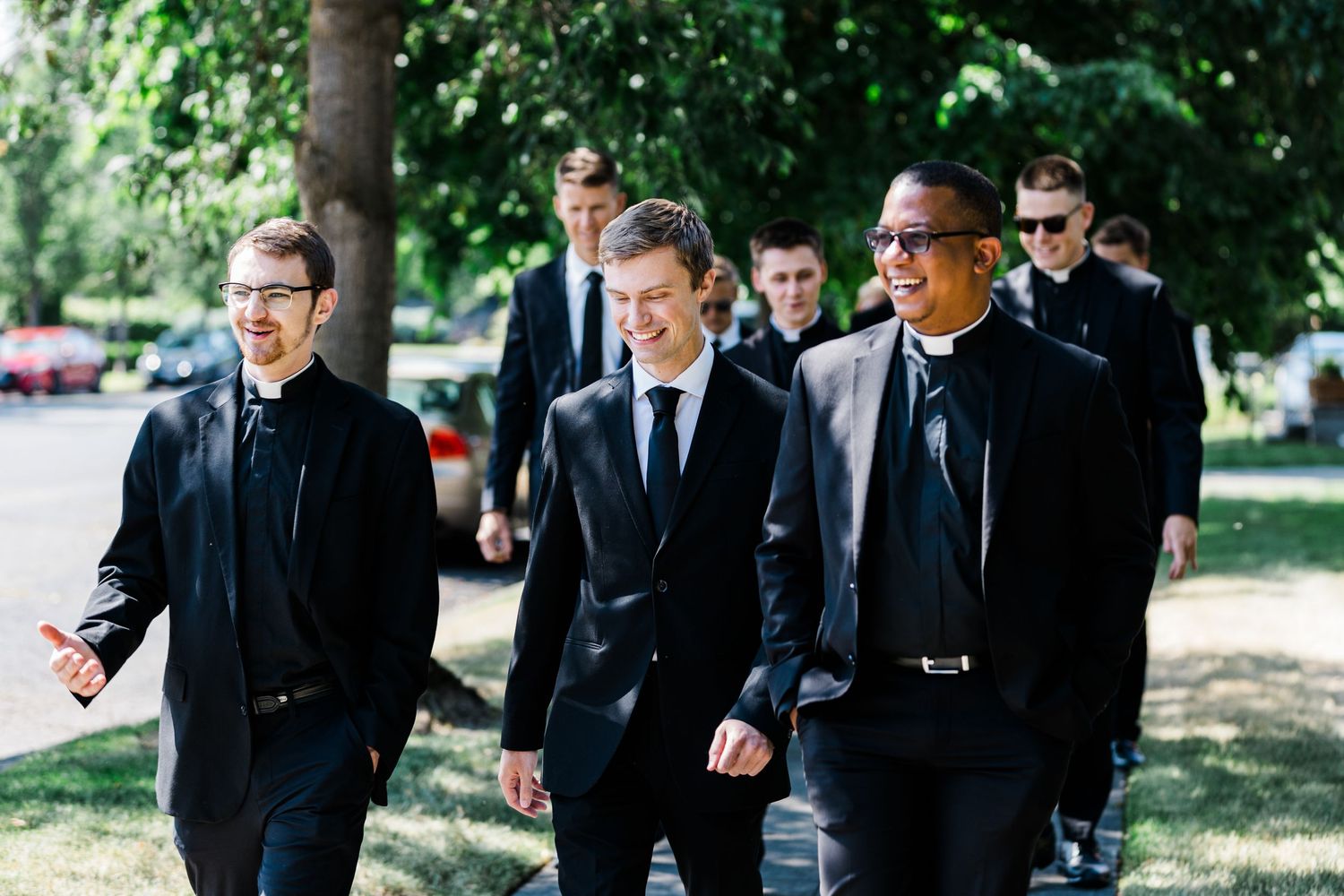 Group of men in black suits walking together outdoors on tree-lined path.