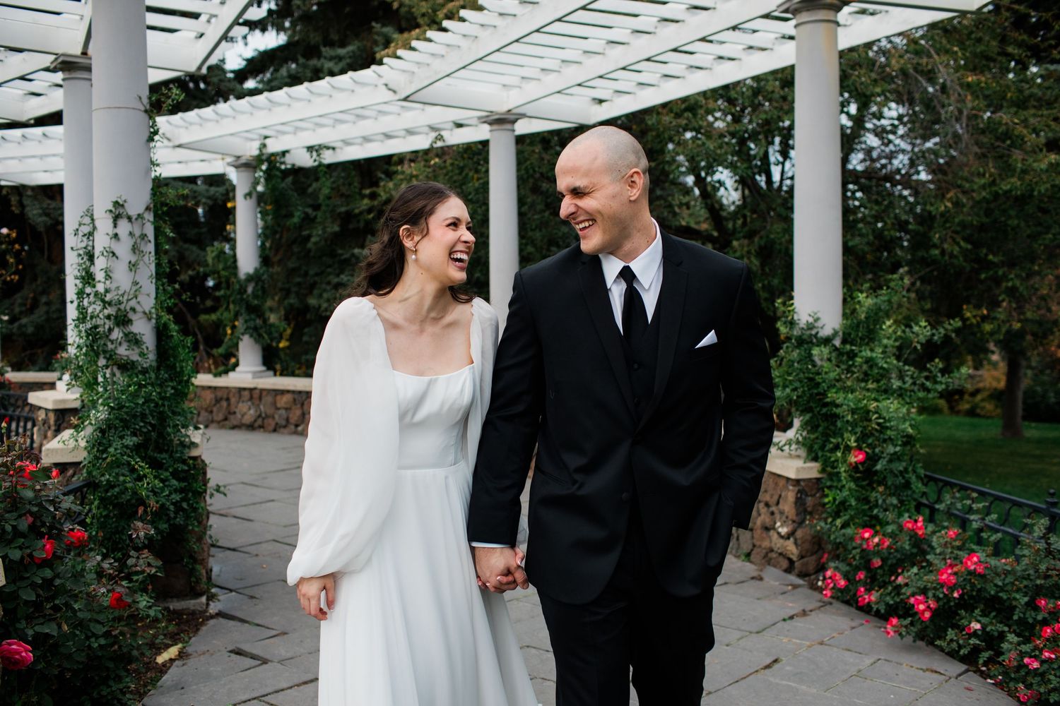Couple poses under white pergola surrounded by blooming roses in garden setting.