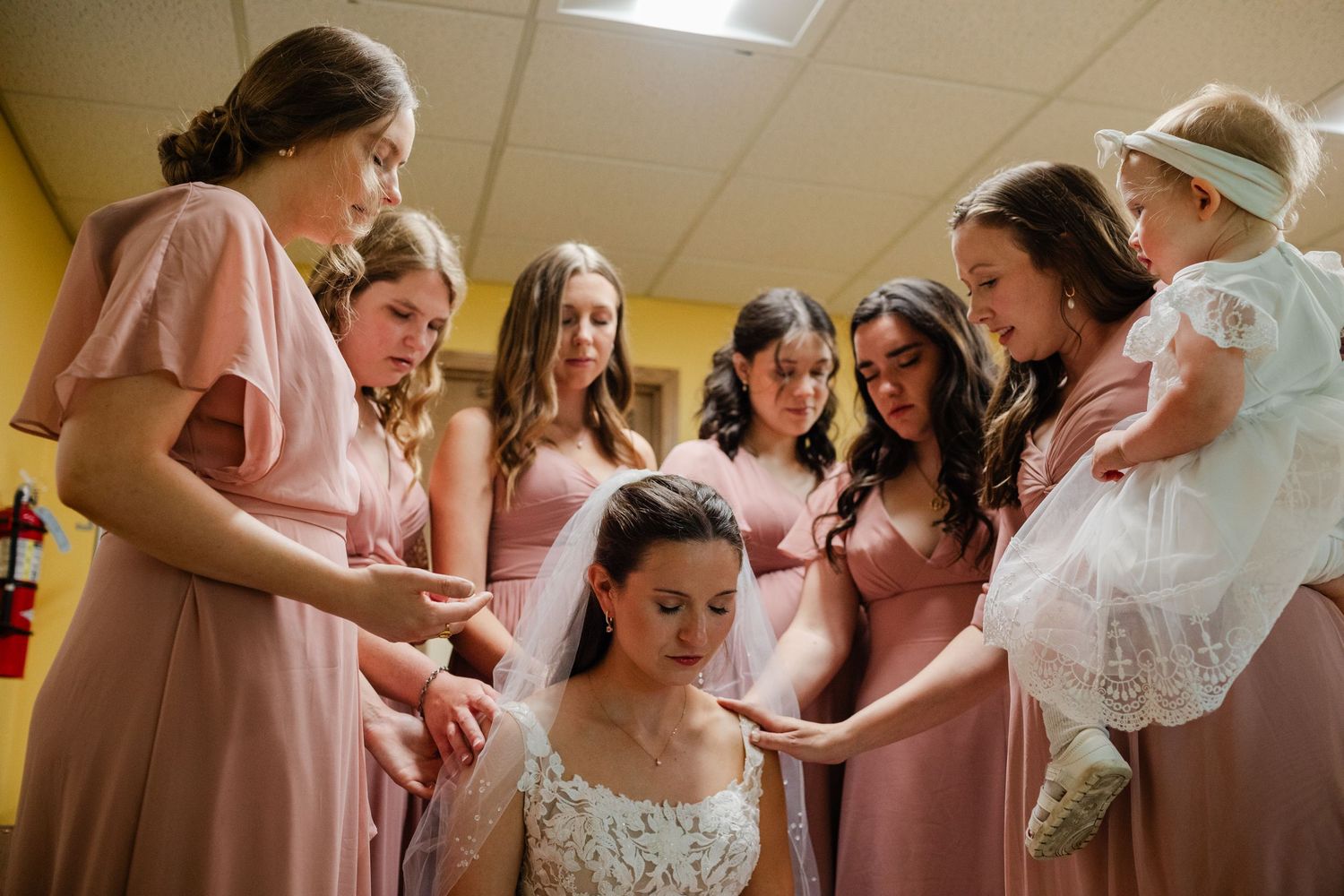 Group gathered in yellow room wearing pink dresses during wedding preparation.