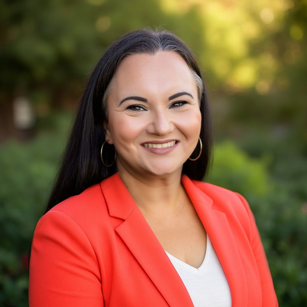 Professional headshot of a person wearing a bright coral blazer against a blurred natural background.