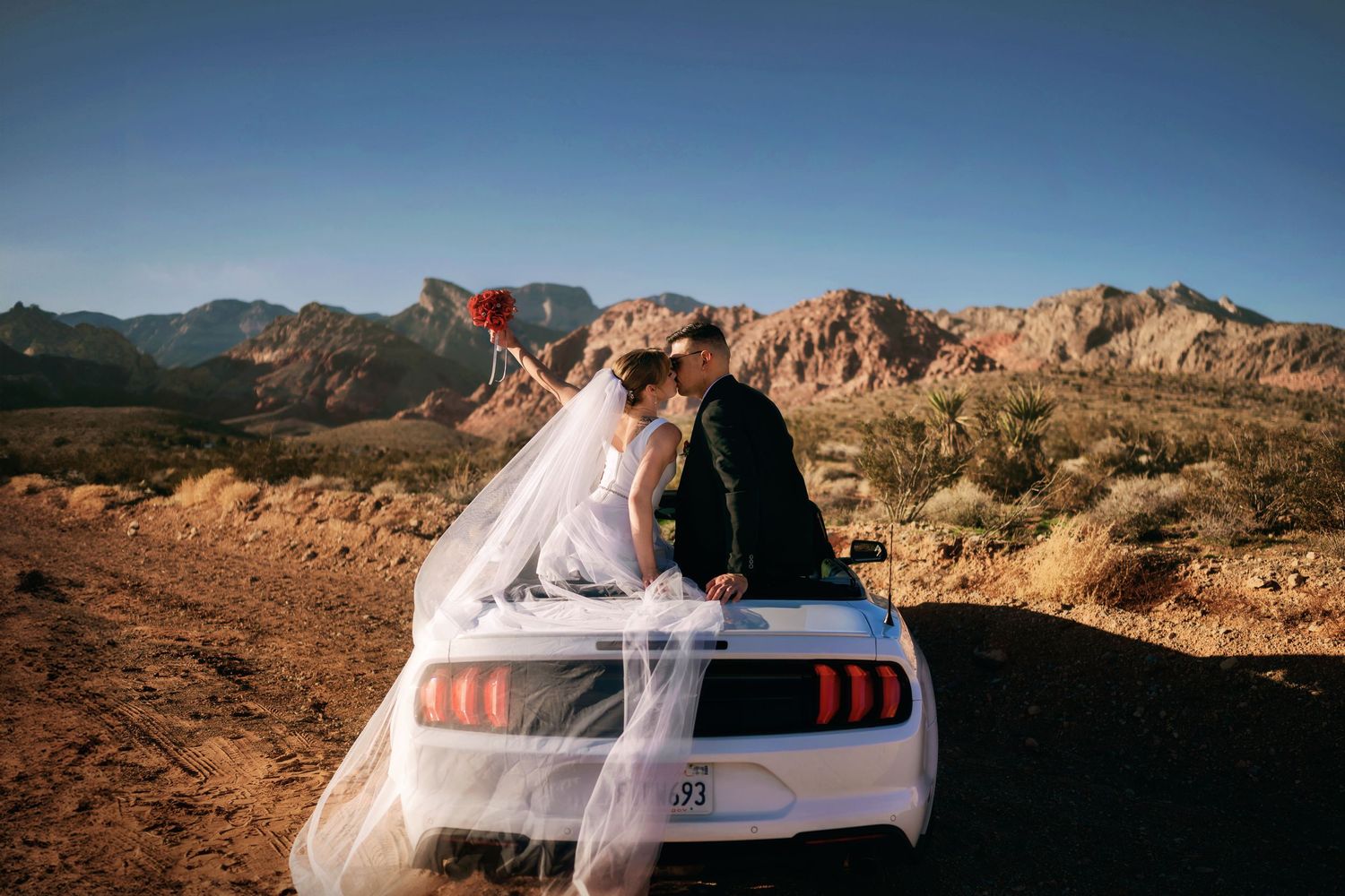 A couple sits on the trunk of a white Ford Mustang during sunset in a scenic desert mountain landscape.