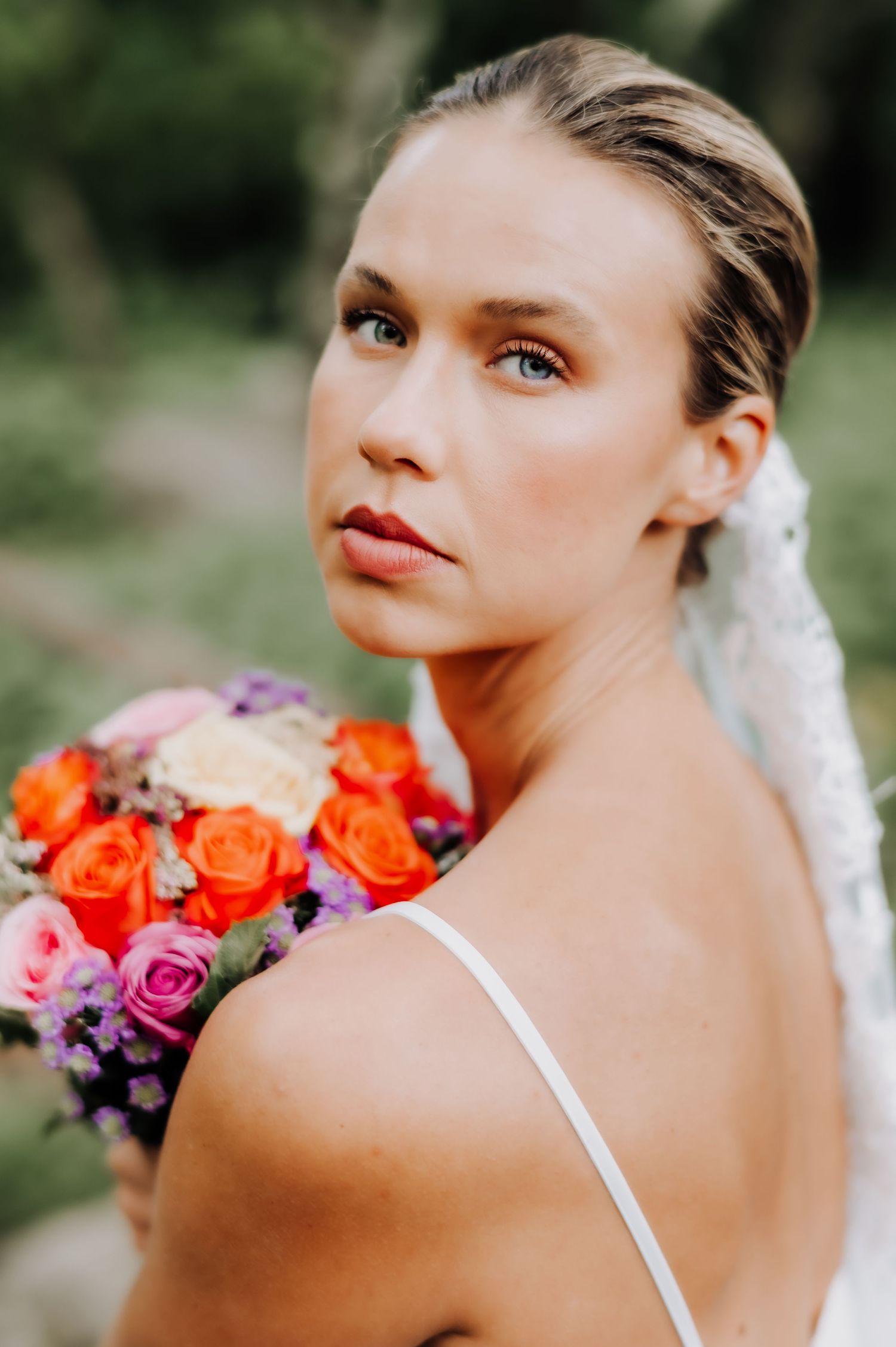 A bride in a white dress strap holds a vibrant bouquet of orange and pink roses during an outdoor photoshoot.