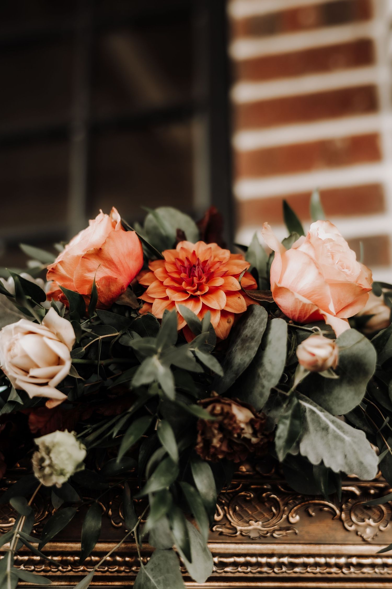 Close up moody photograph of a floral arrangement with peach roses, orange dahlia, and green foliage on an ornate surface.