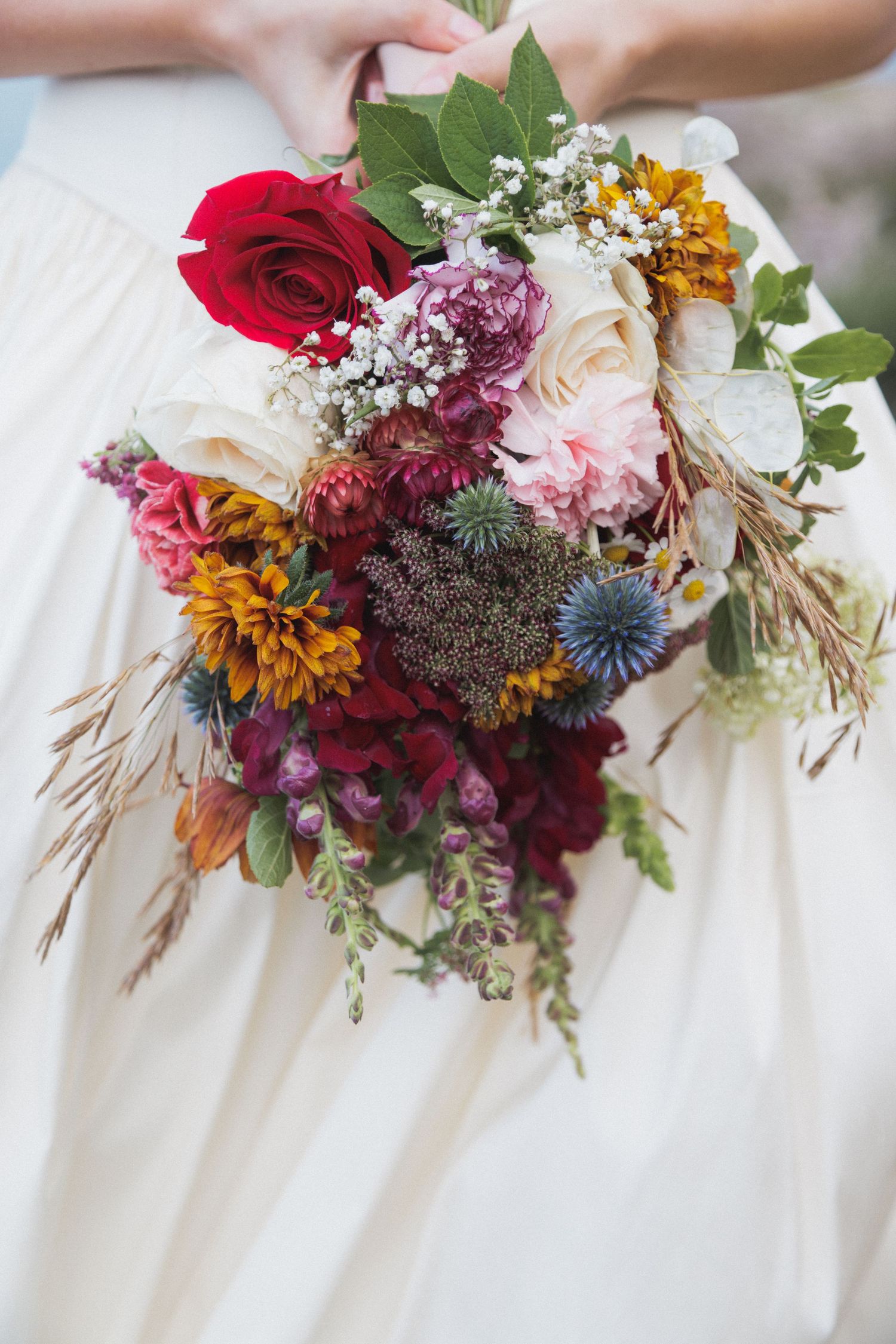 Four bridesmaids hold coordinating fall wedding bouquets with burgundy roses, sunflowers, and wildflowers against white dresses.