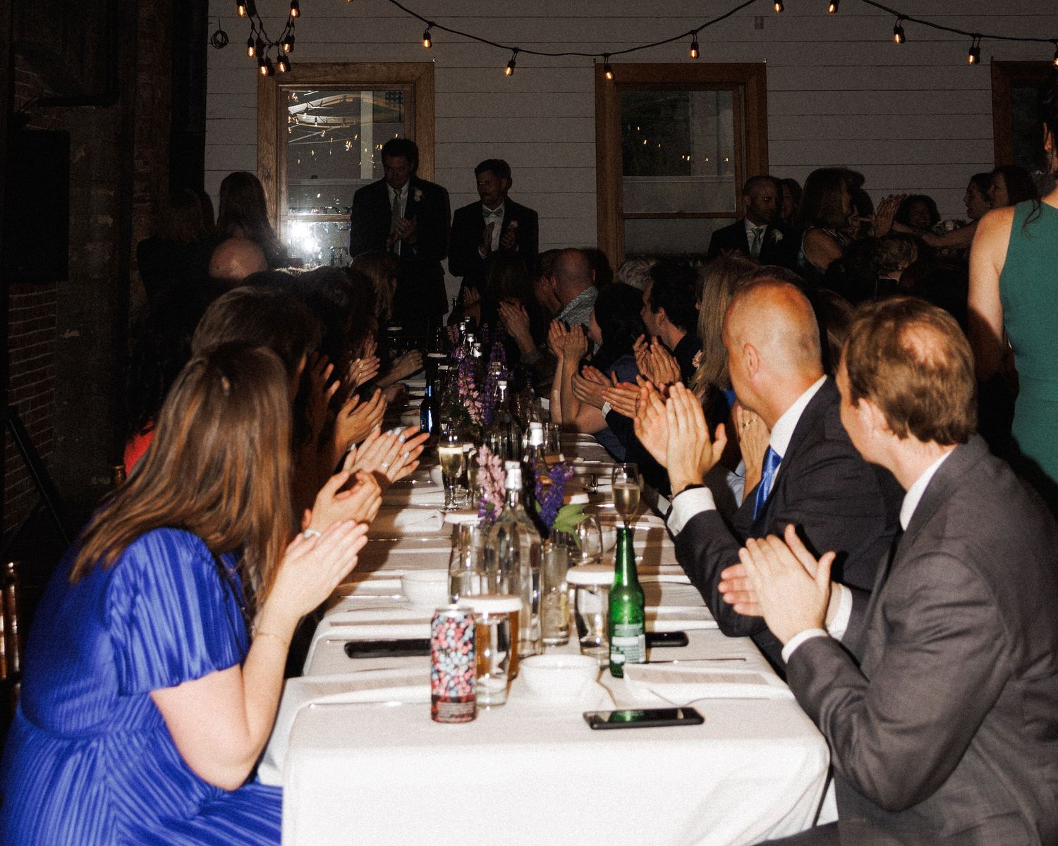 Guests applaud at a long table under string lights at The Well Room, by Kaitlyn Yvette, Newfoundland wedding photographer.