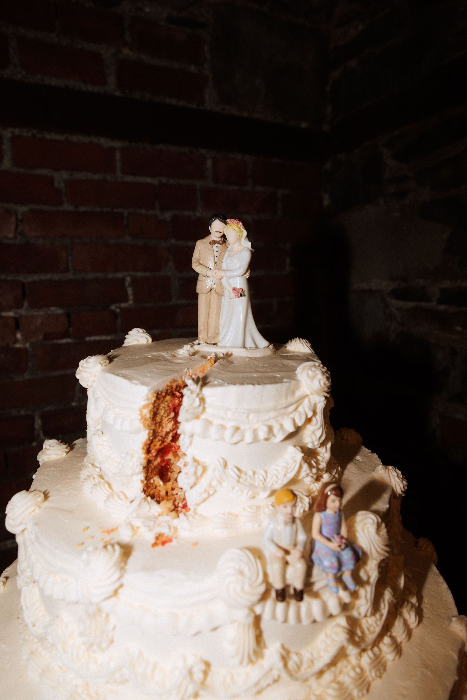 A damaged white wedding cake with traditional bride and groom cake topper against a brick wall background.