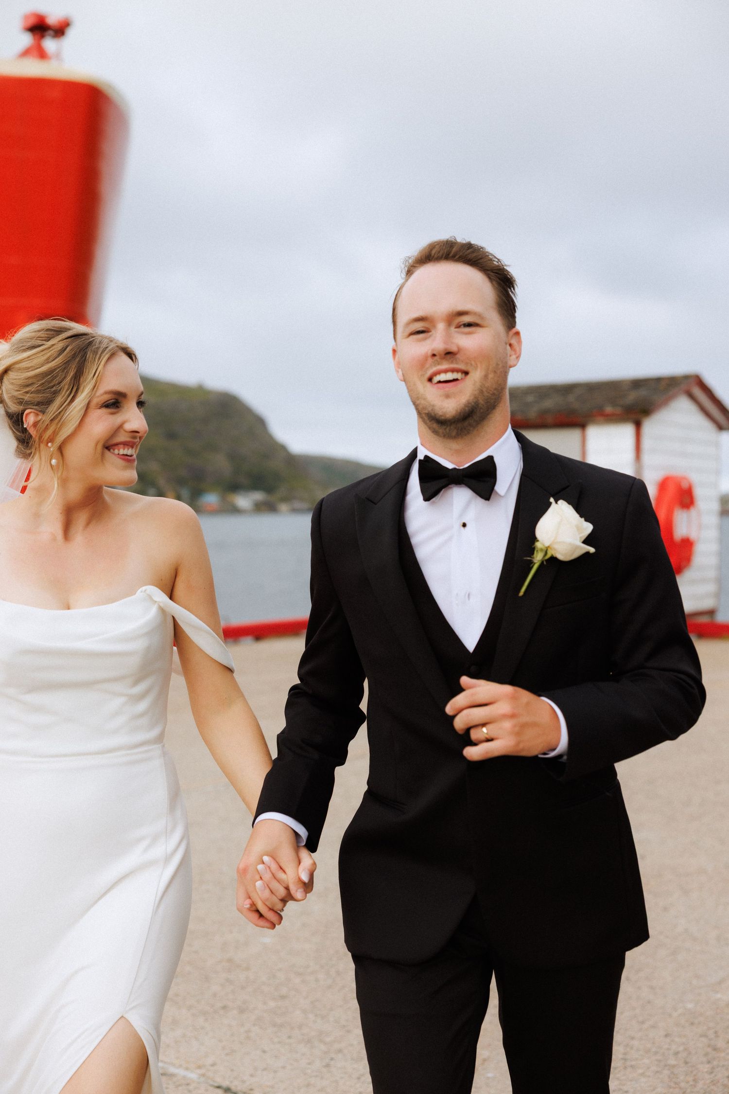 A happy couple in wedding attire laugh while walking together on a beach with red posts in the background.