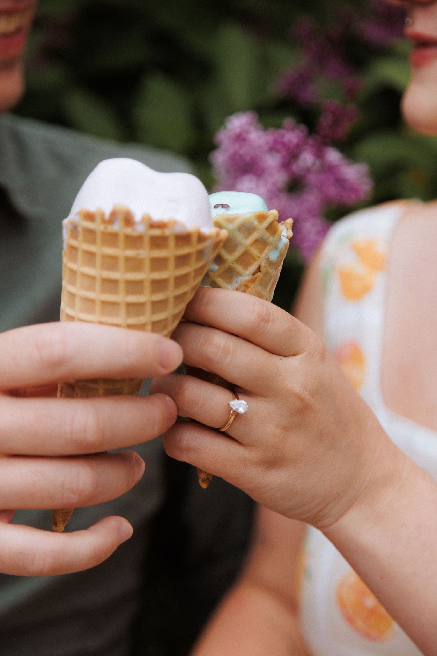 Two hands holding ice cream cones with waffle cones against a blurred floral background, one hand wearing a ring.