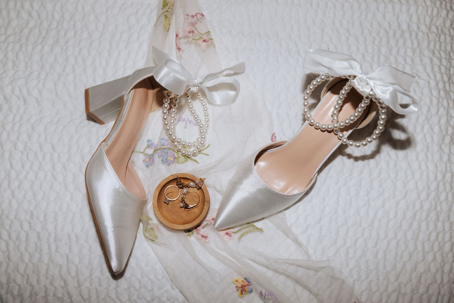 Elegant white bridal shoes with pearls and a delicate bow accent displayed with wedding rings on white textured fabric.