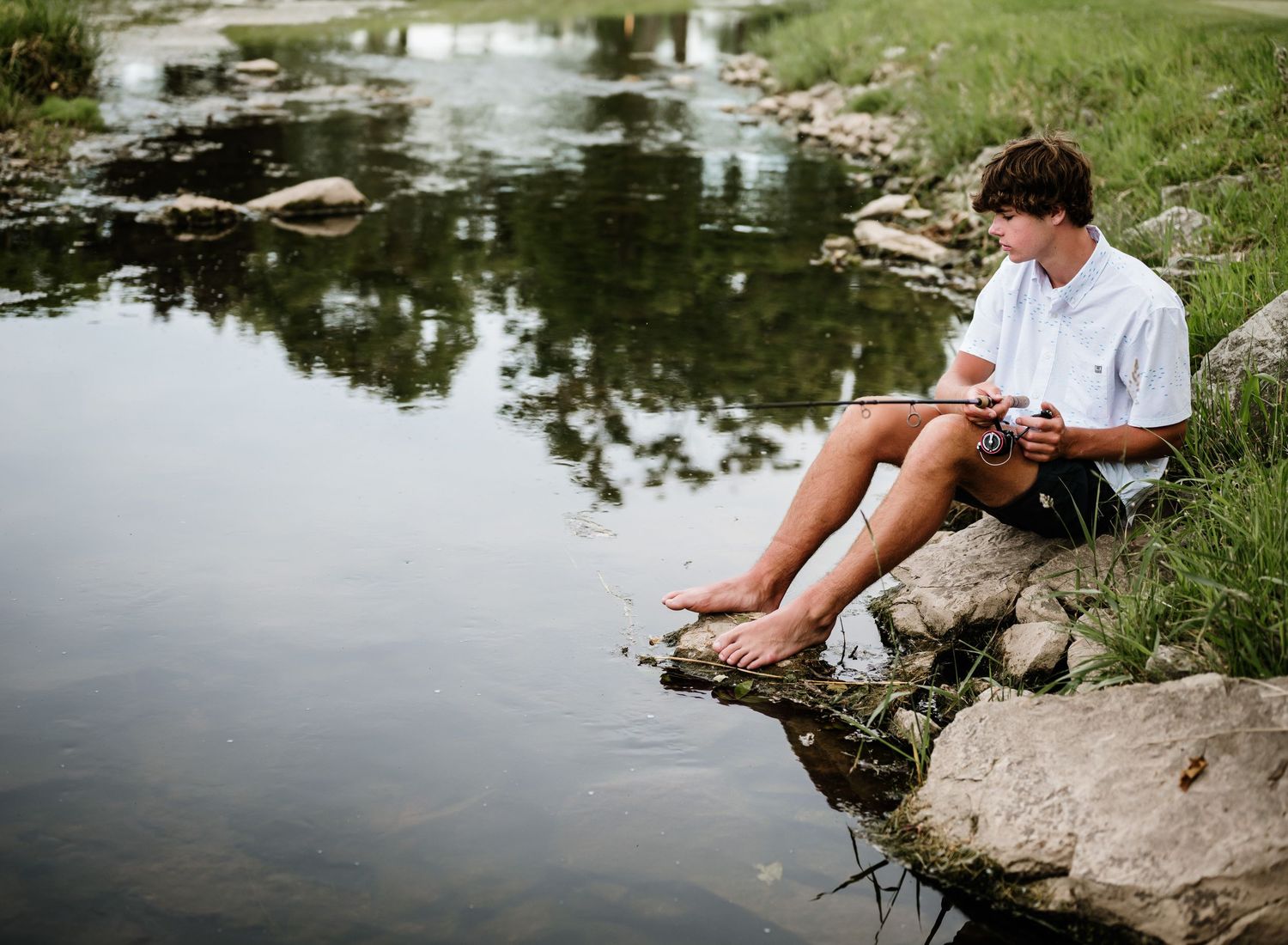 Someone in a white shirt sits on a rock by a peaceful pond while looking down at a mobile device.