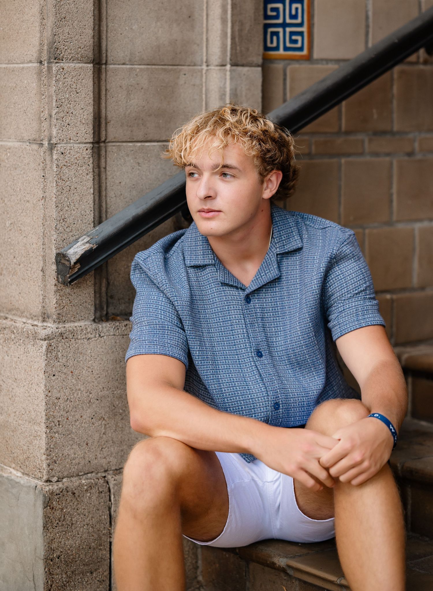Casual senior portrait of someone in blue polo shirt sitting on stone steps near architectural details.