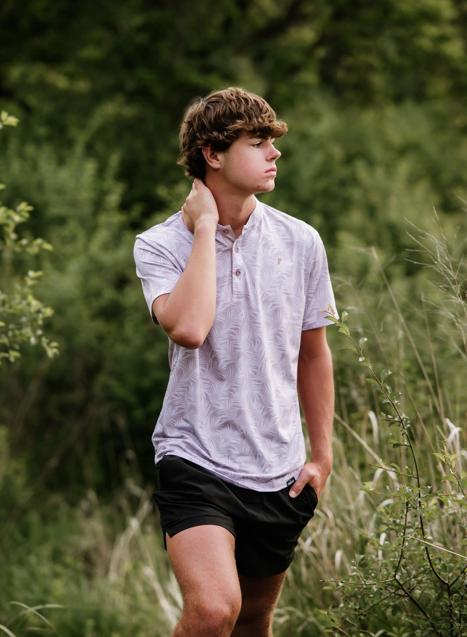 Young person in white polo shirt and dark shorts poses thoughtfully in a lush green outdoor setting.
