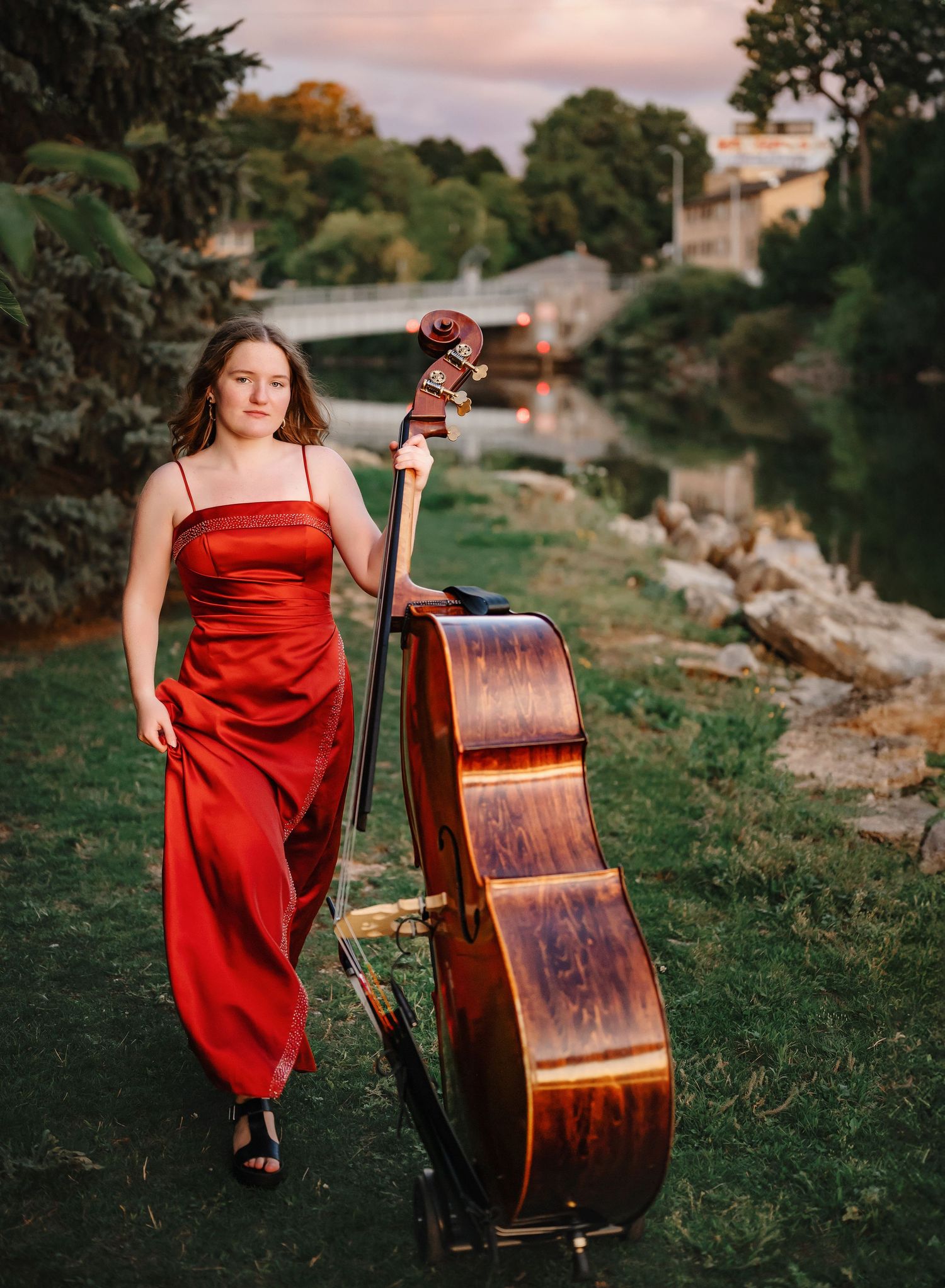 Senior in a flowing red dress poses outdoors with an upright bass against a scenic natural backdrop.