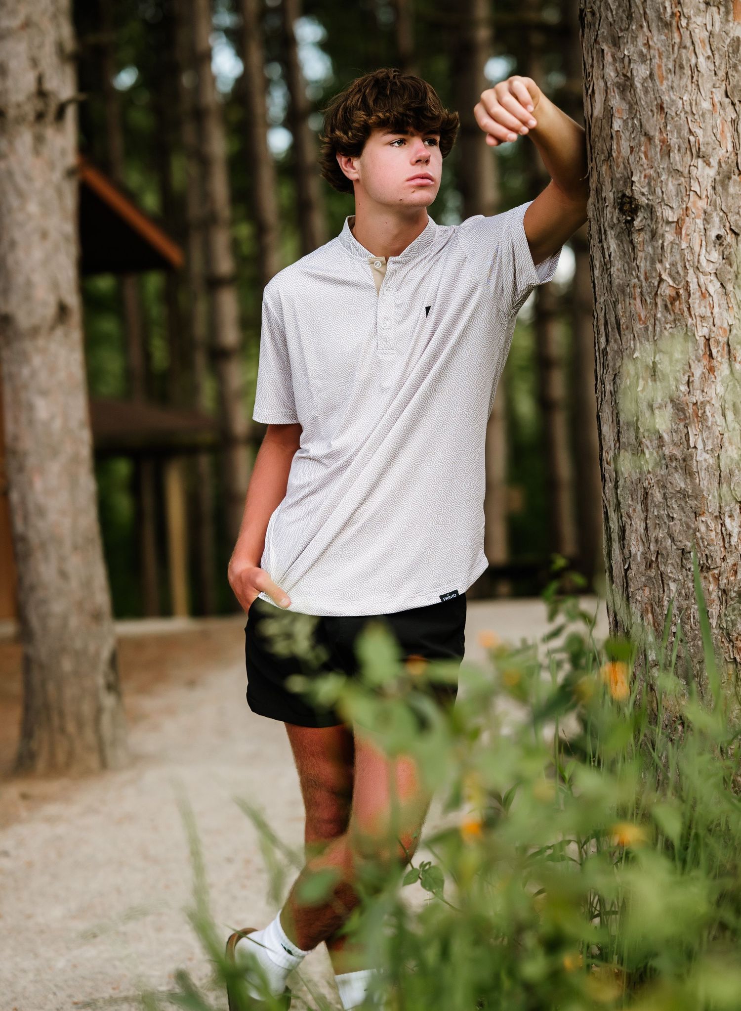 Person in white polo shirt and brown shorts leans against tall pine trees in an outdoor forest setting.