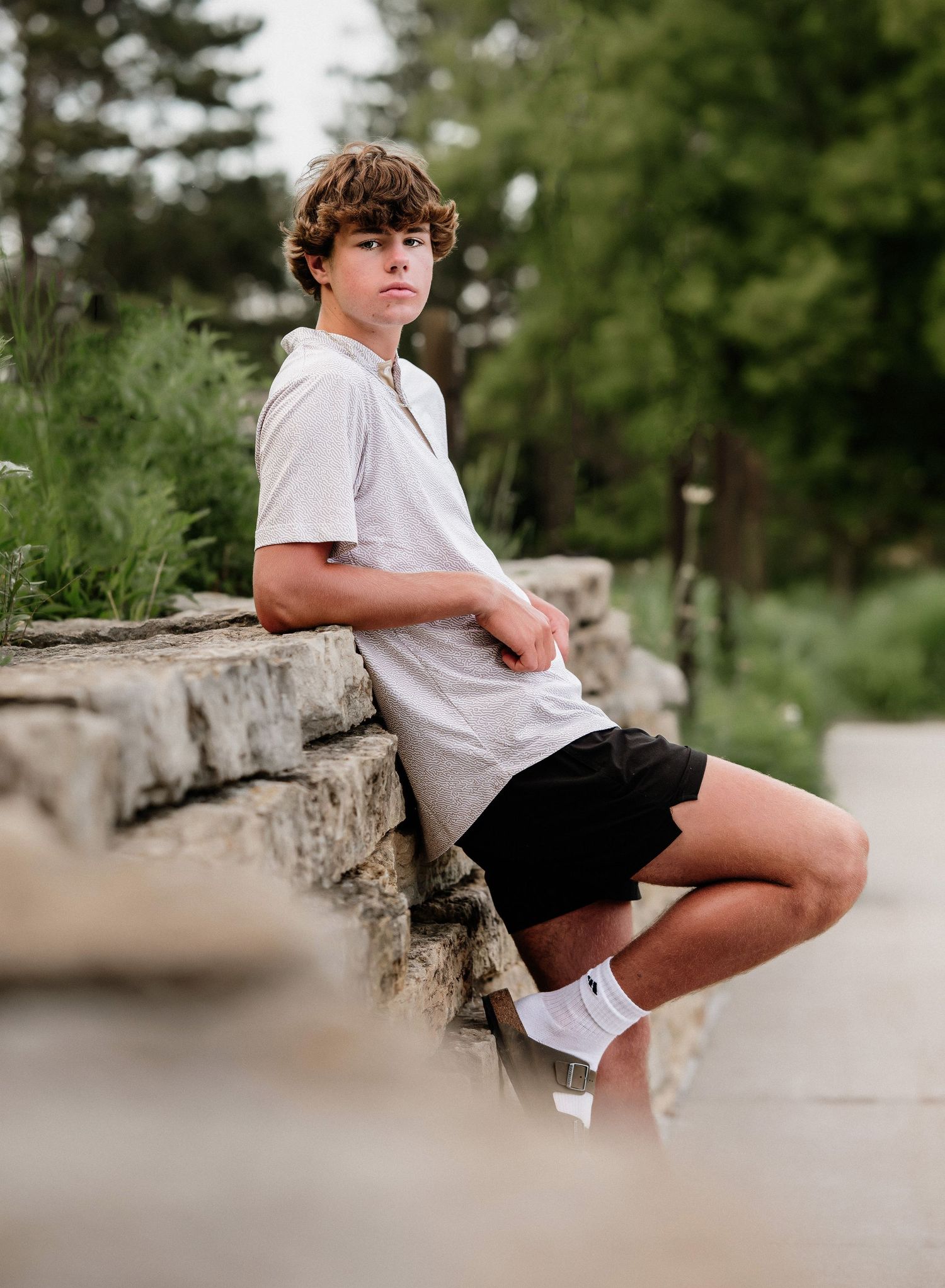 Person in white t-shirt and black shorts sits casually on stone wall in outdoor park setting.