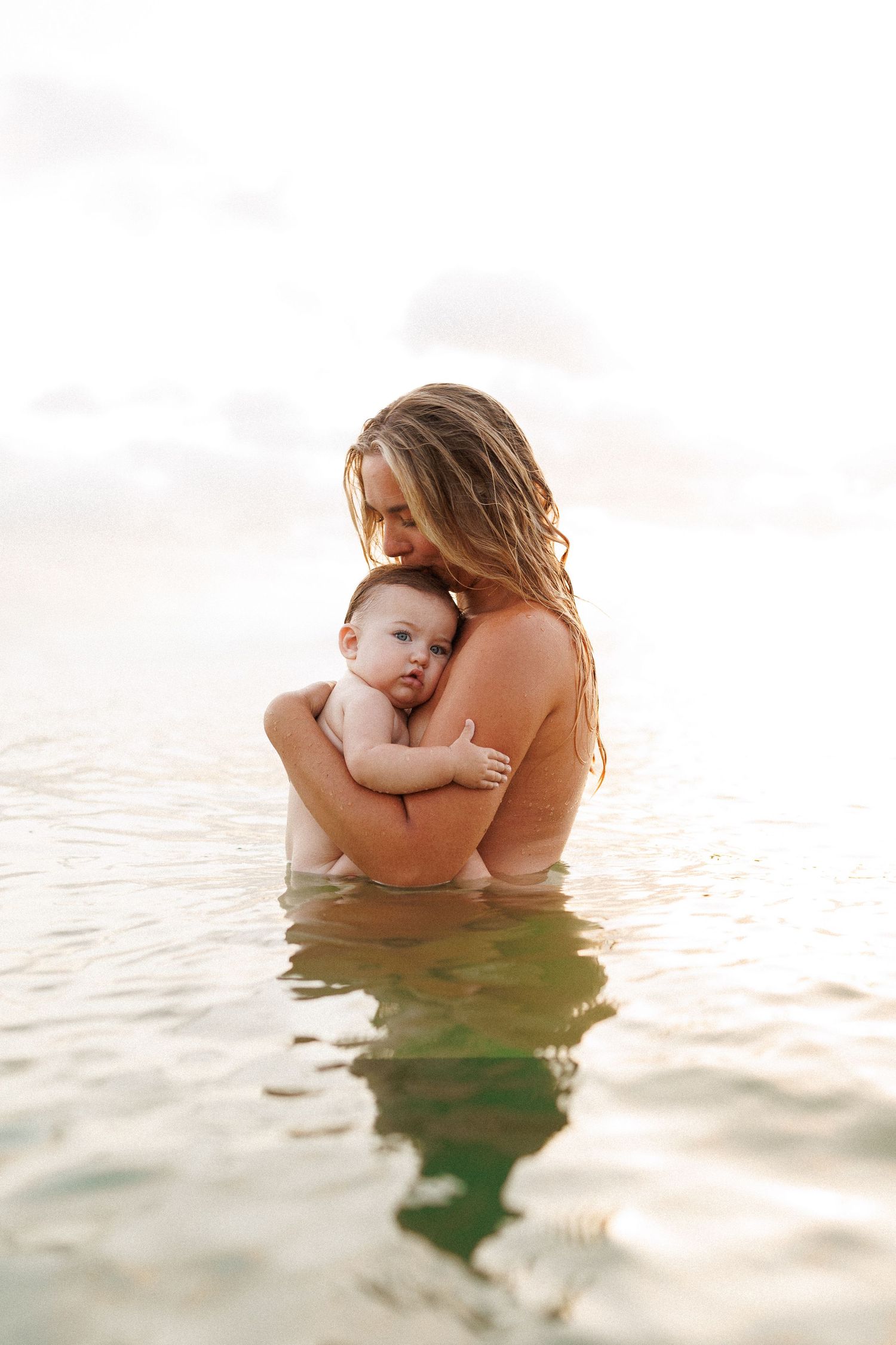 A tender moment captured as mother and baby share an intimate embrace while standing in tranquil ocean waters at sunset.