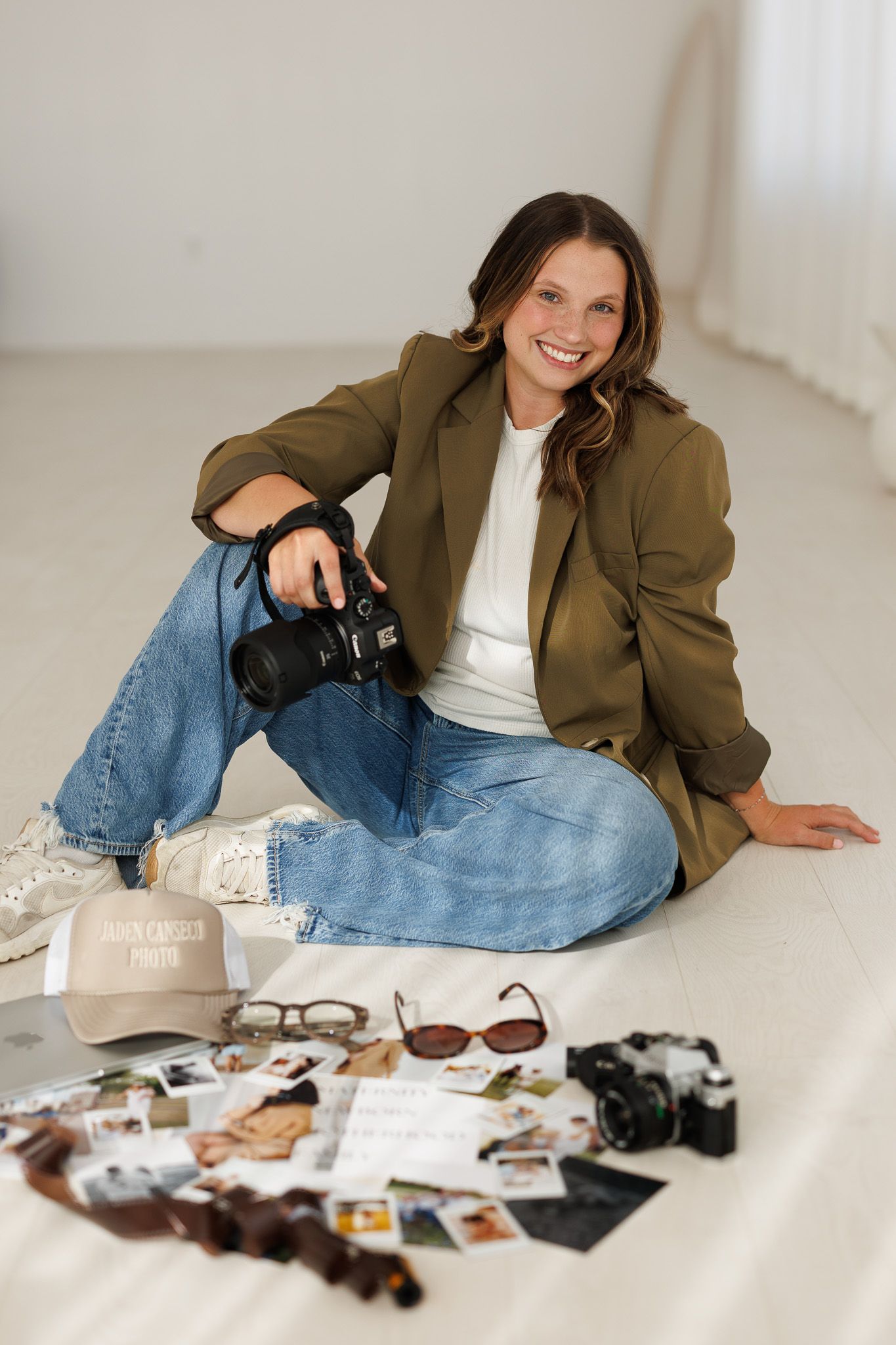 Professional photographer sitting on floor with camera gear, wearing olive blazer, white top and jeans, surrounded by equipment.