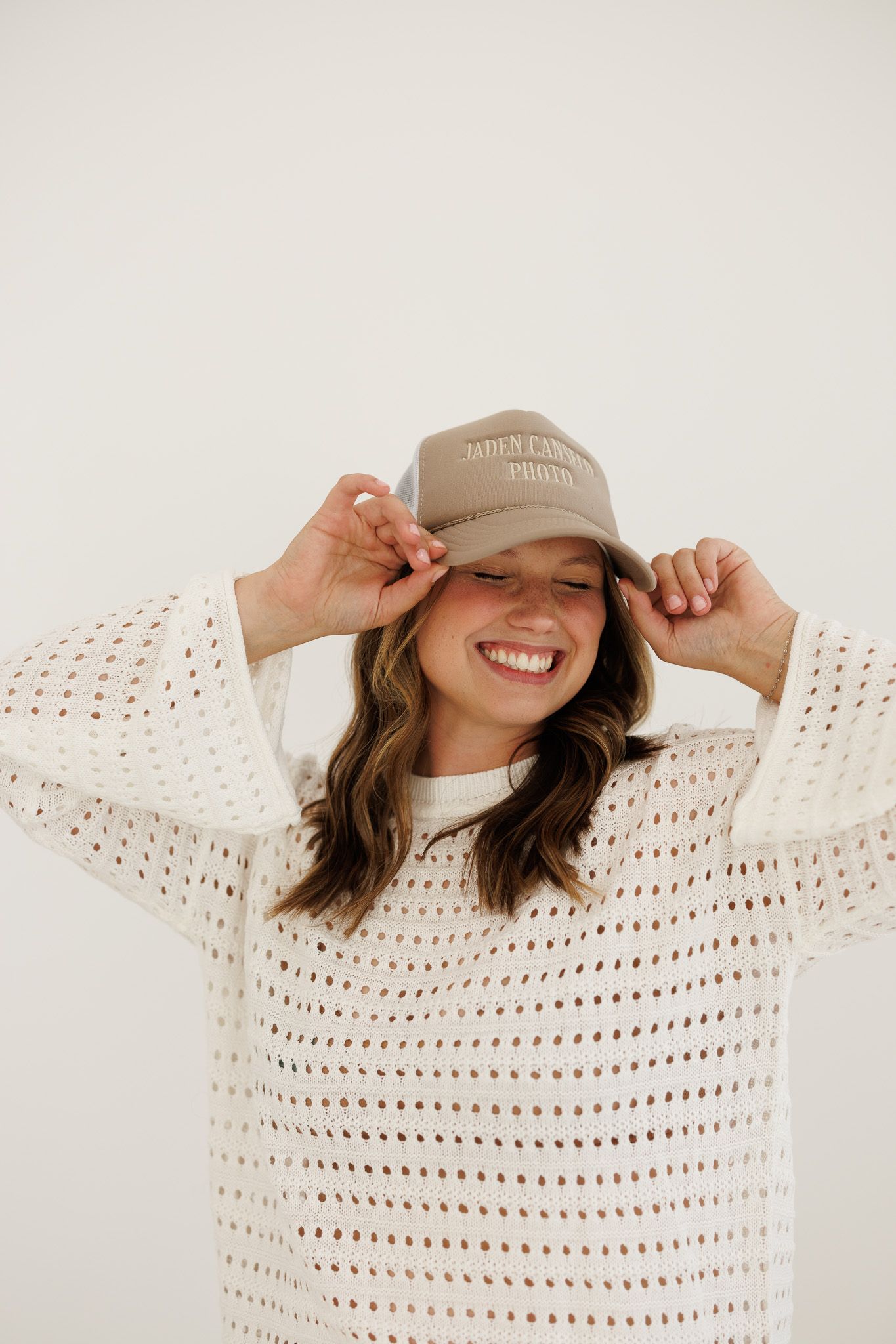 Smiling person in a cozy white knit sweater and beige baseball cap playfully adjusting their hat while posing.