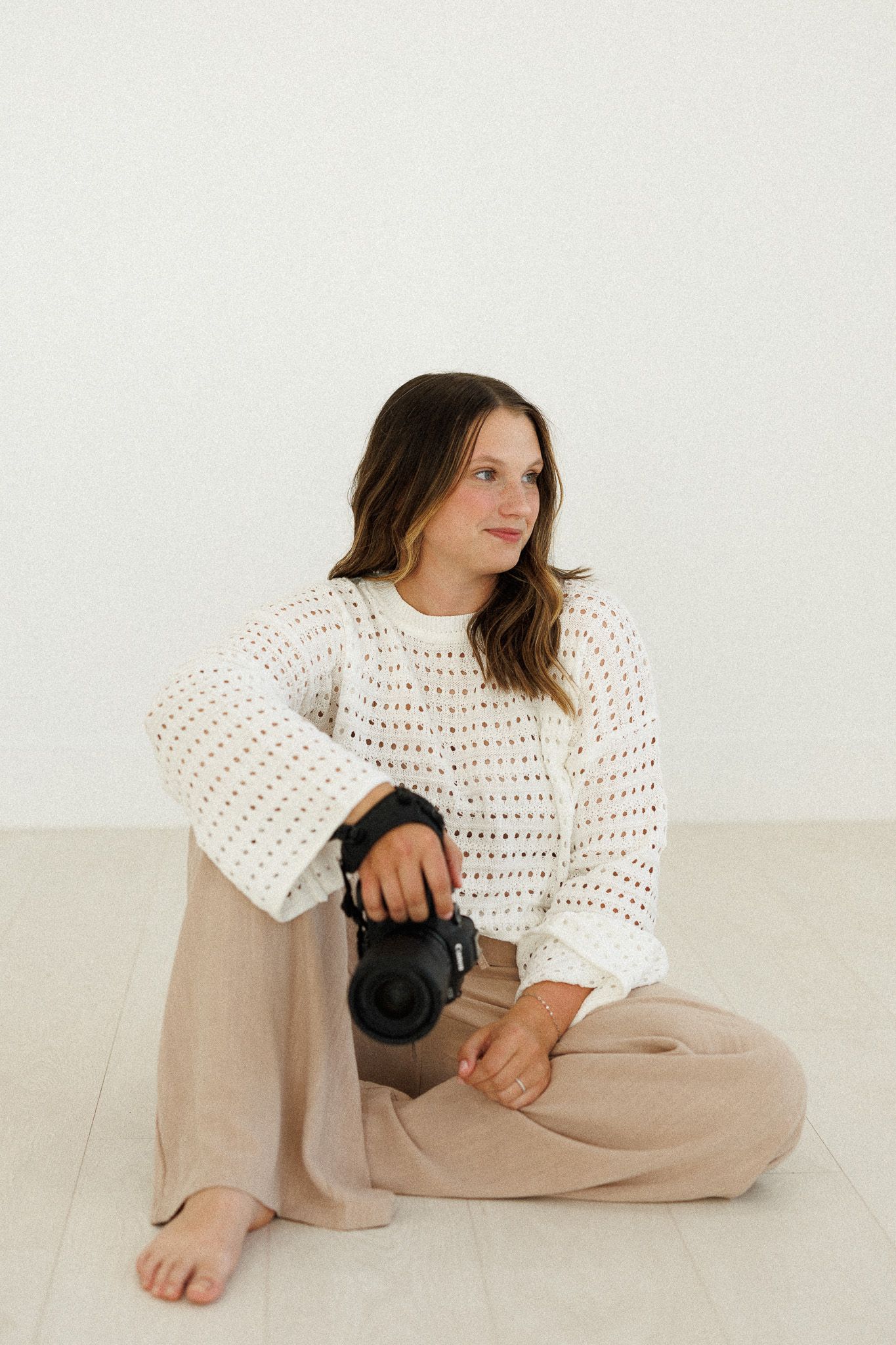 Person wearing white crochet sweater and beige pants sits casually on floor with camera in minimalist setting.
