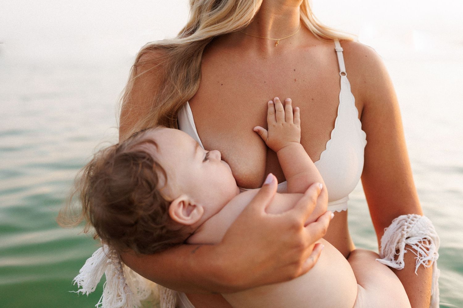 A tender breastfeeding moment captured near calm water with mother wearing white and baby in soft lighting.