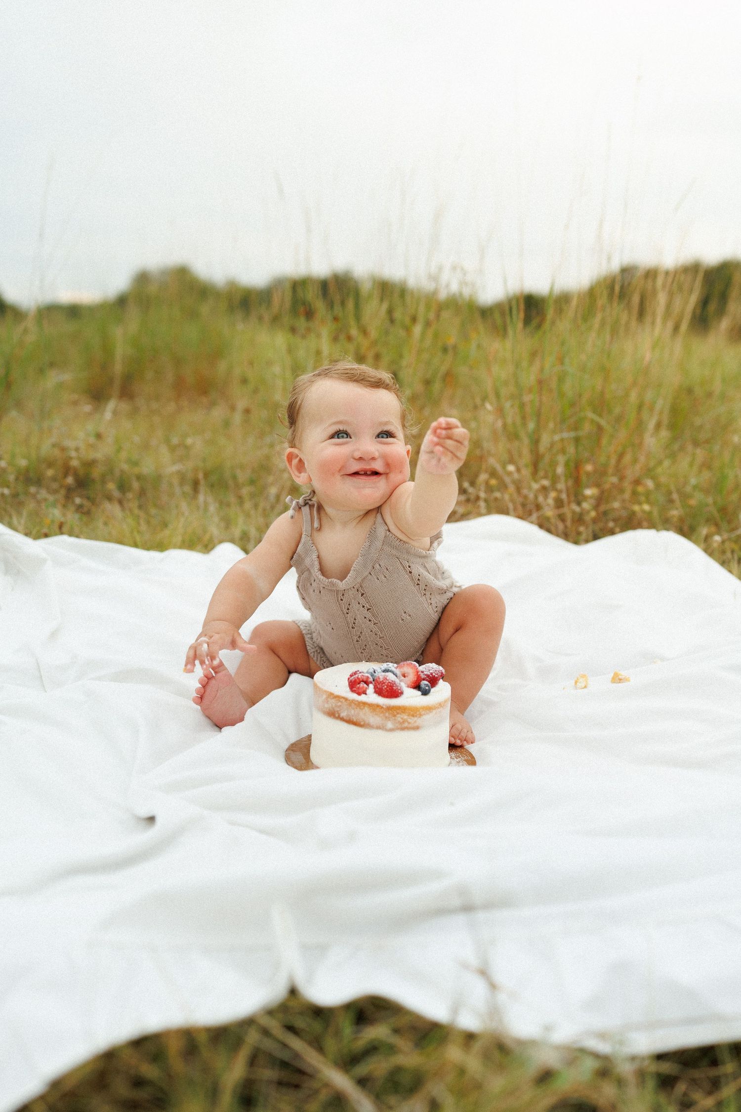 A baby wearing gray lies on a white blanket in a grassy field, enjoying a smash cake dessert.