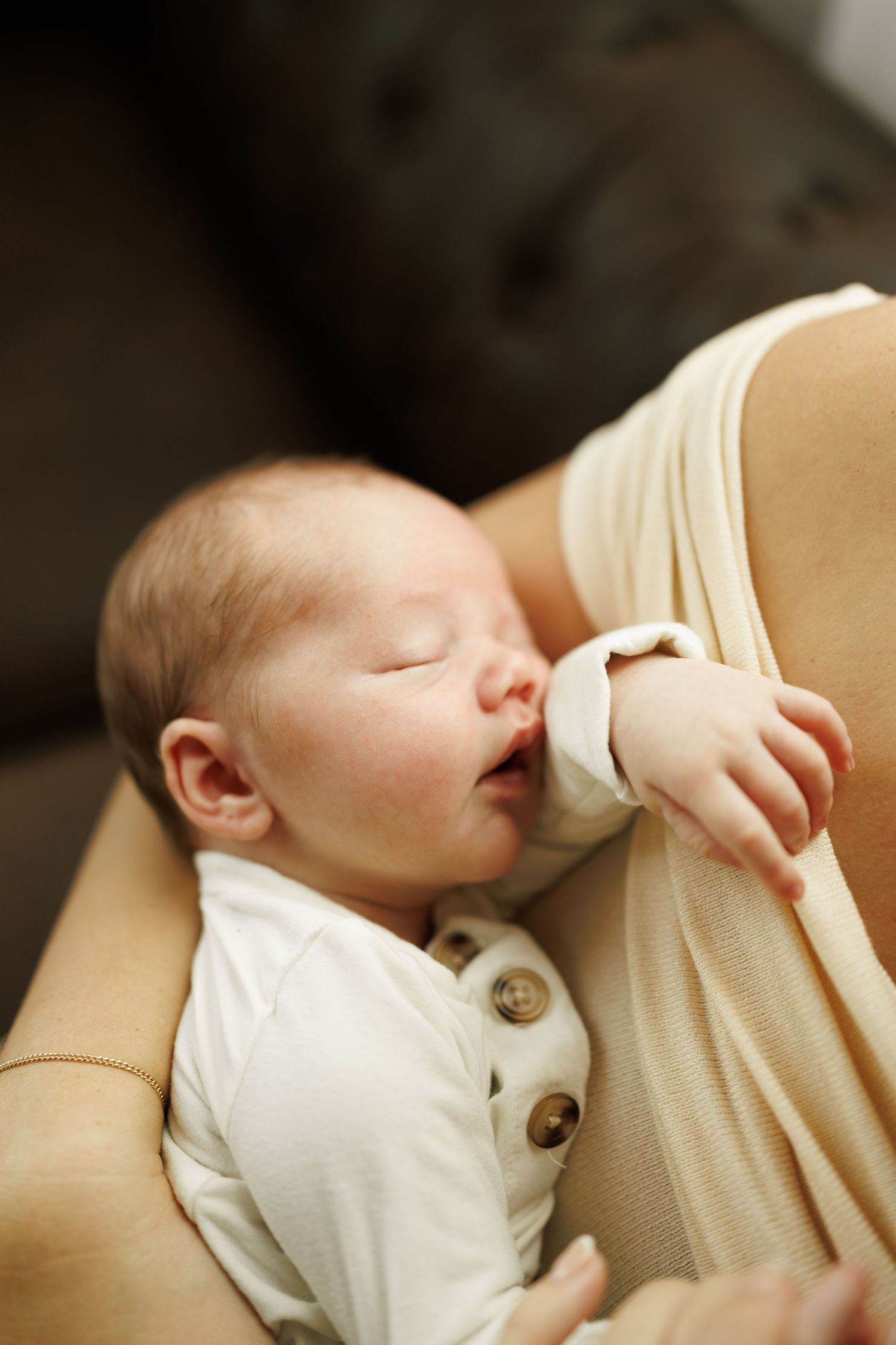 A baby in white sleeps peacefully while breastfeeding against a beige fabric background.