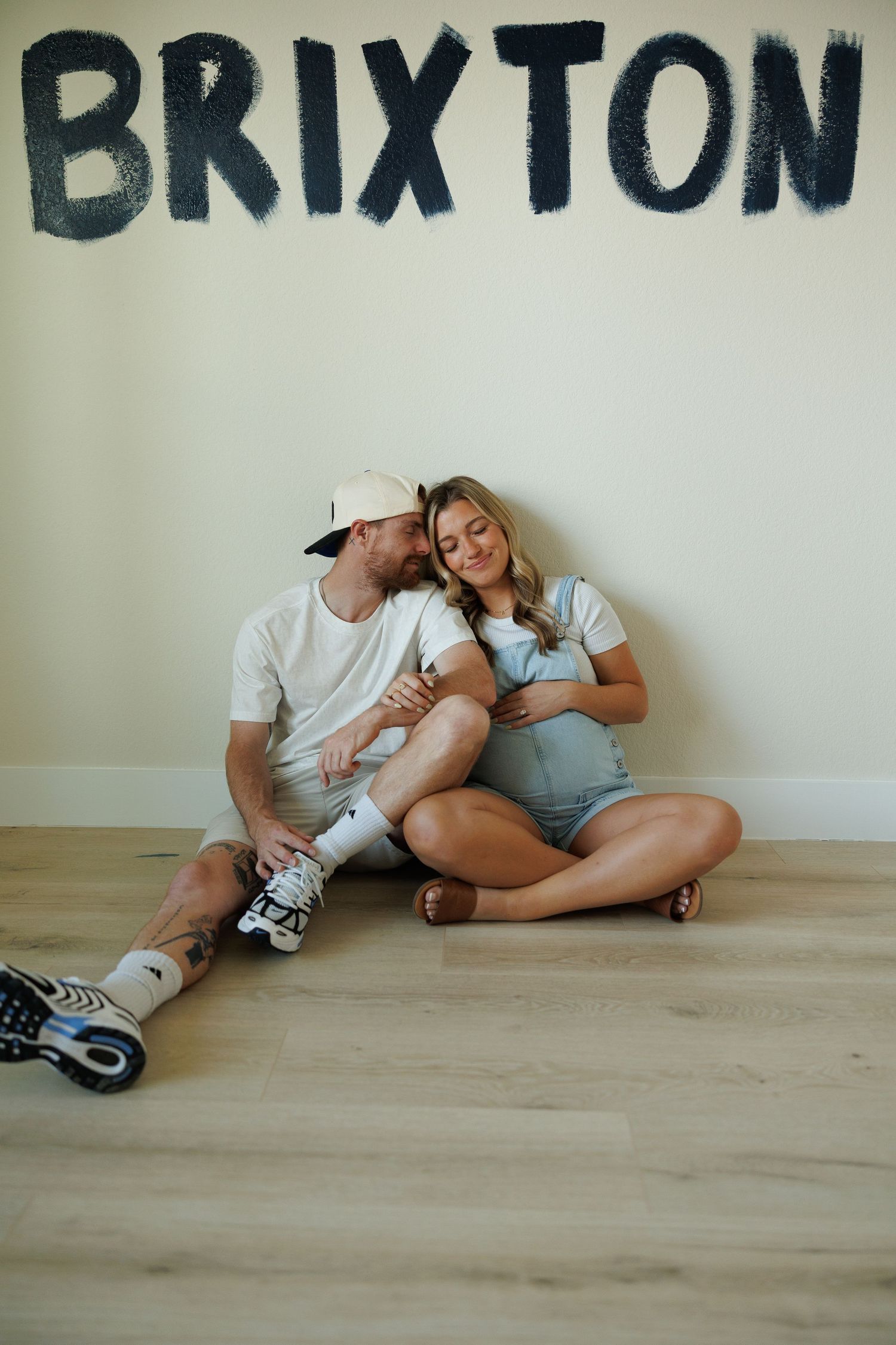 A couple sits together on wooden floor beneath black Brixton text on wall wearing casual white t-shirts.