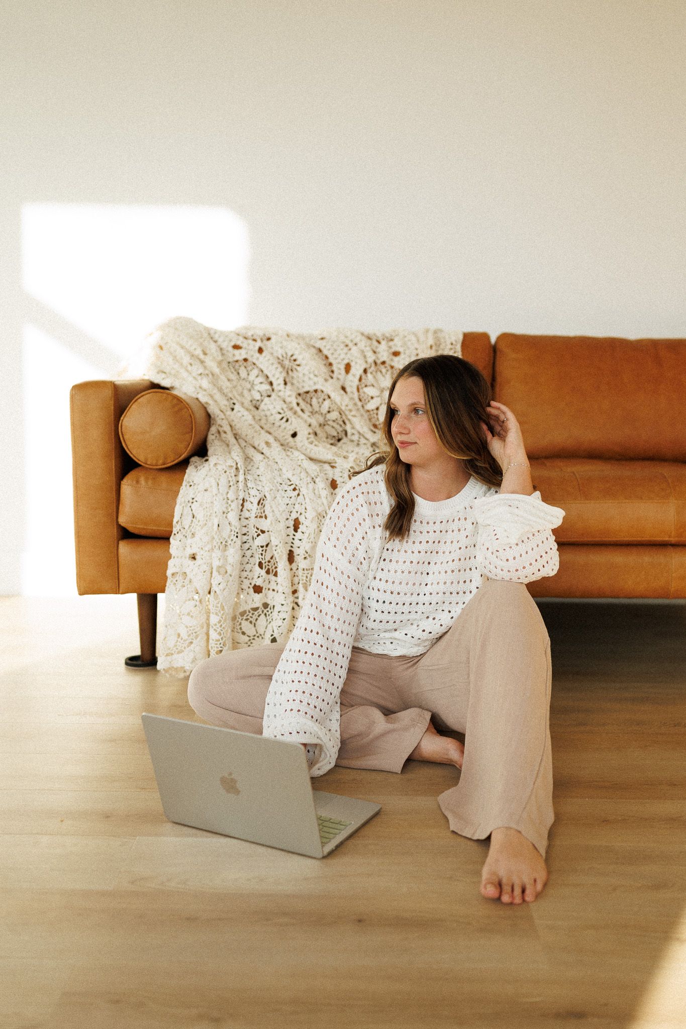 Someone in a cozy white sweater works on laptop while sitting on hardwood floor beside tan leather sofa.