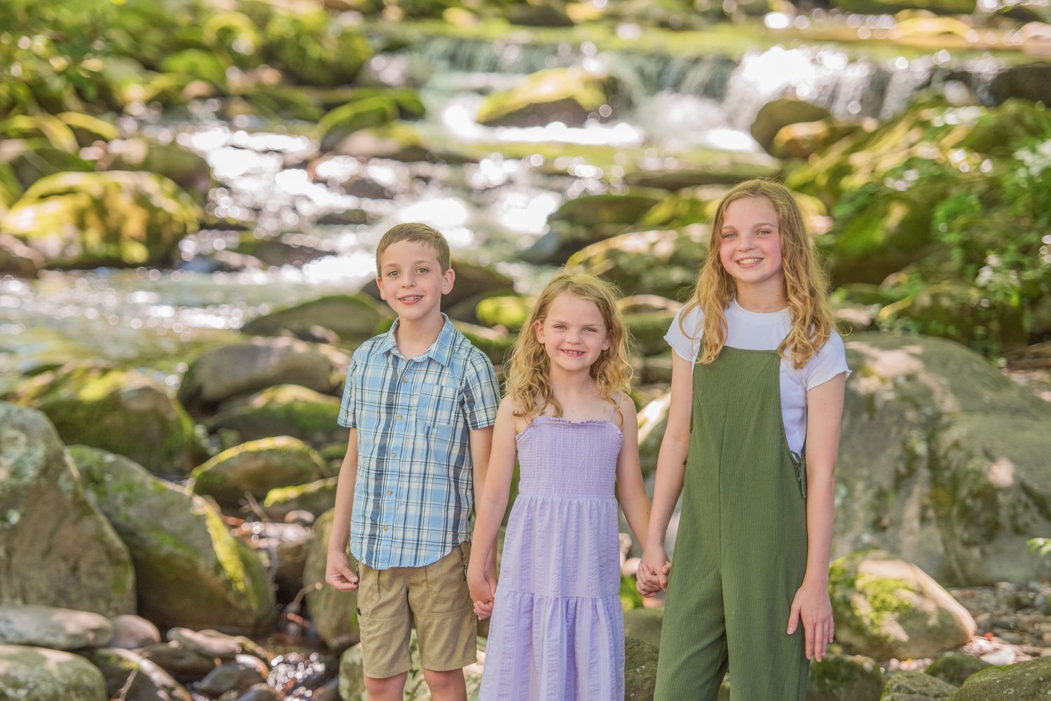 Three children smile while standing together near a rocky stream in a lush outdoor setting by family portrait Gatlinburg photographer.
