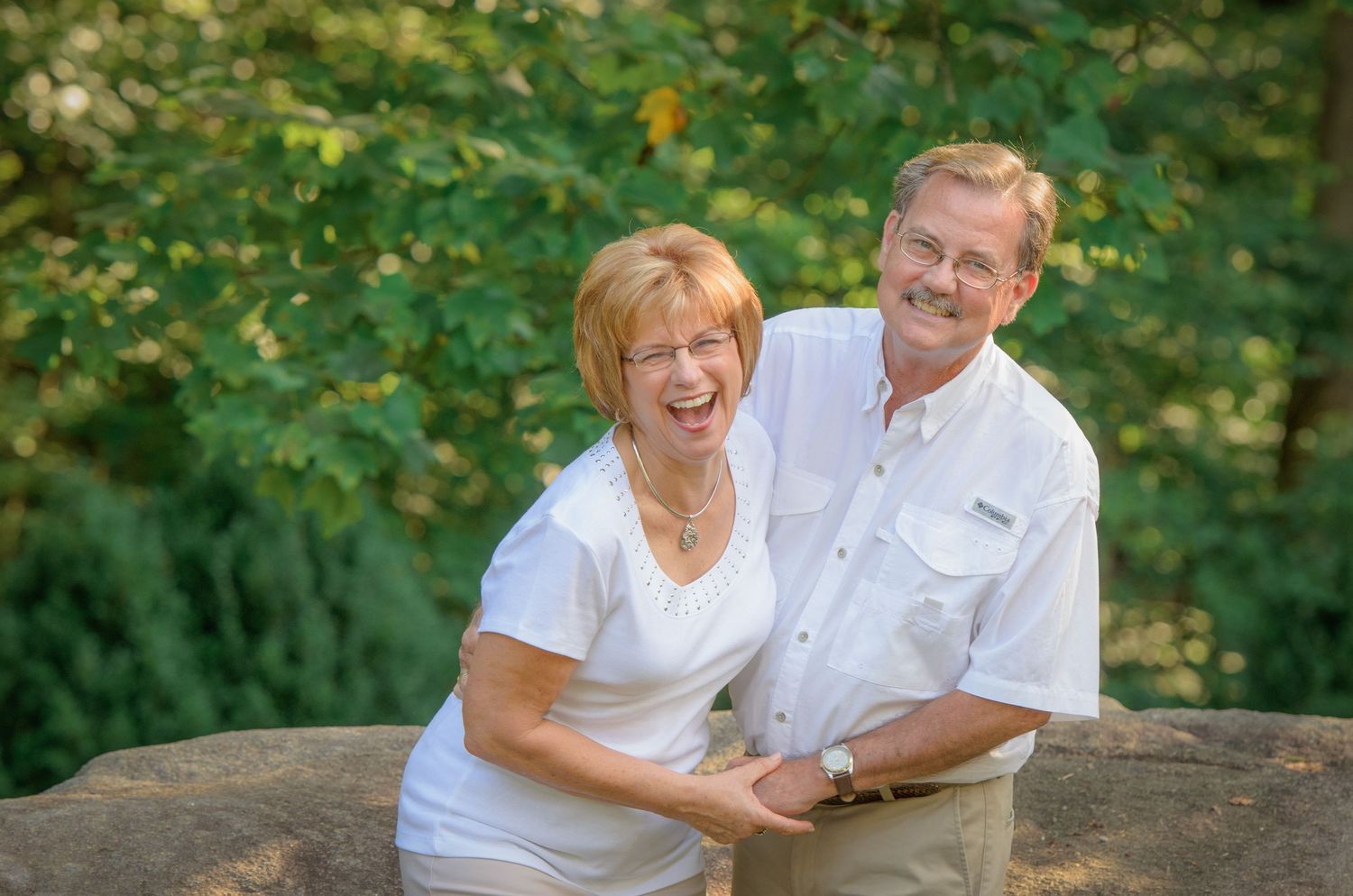 Happy couple in white attire smiling together while leaning on large rock in lush outdoor setting by Gatlinburg TN Photographers.