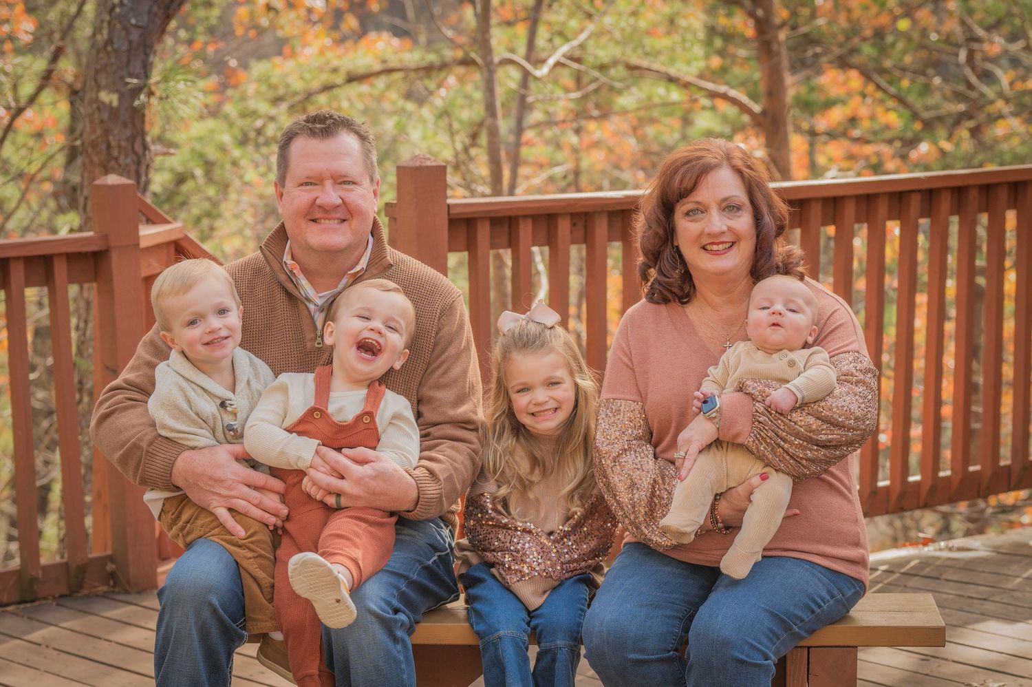 A family with four young children sits together on a wooden deck during autumn with colorful fall foliage in the background at a cabin.