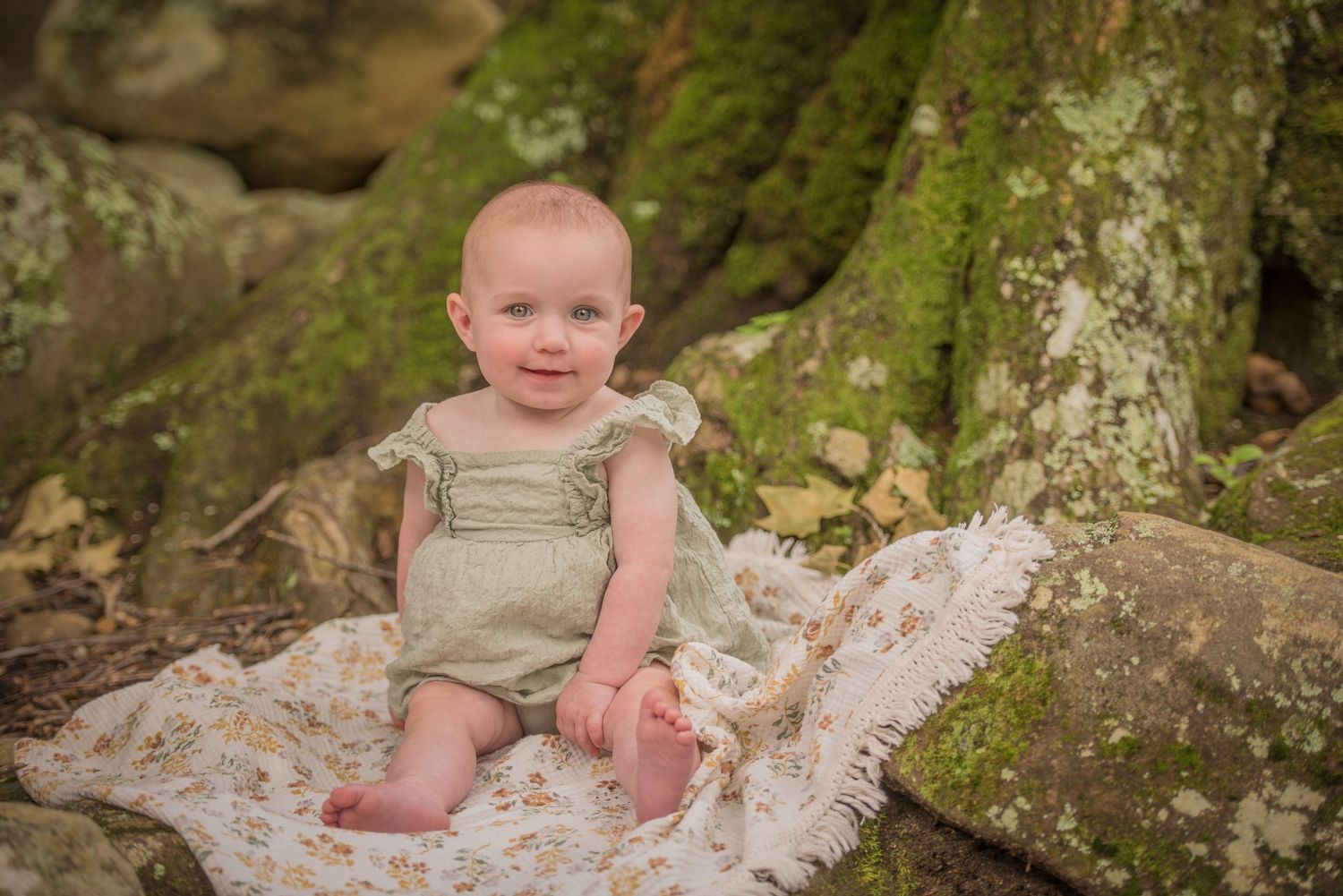 Small child in sage green romper sits on mossy rocks with lace fabric during outdoor nature portrait session in Pigeon Forge TN.